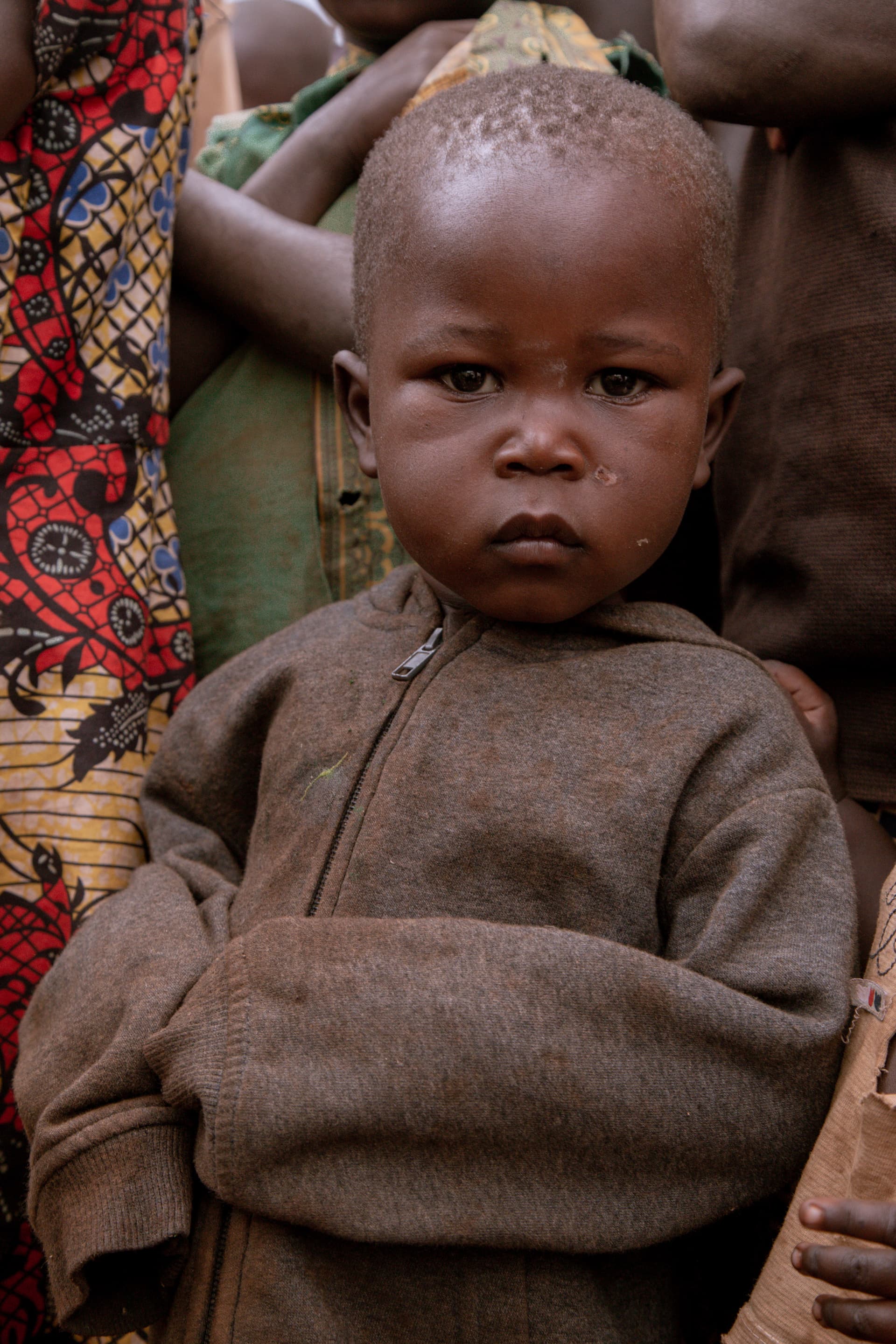 A toddler in a brown sweater stands close to an adult with a serious expression, outdoors.