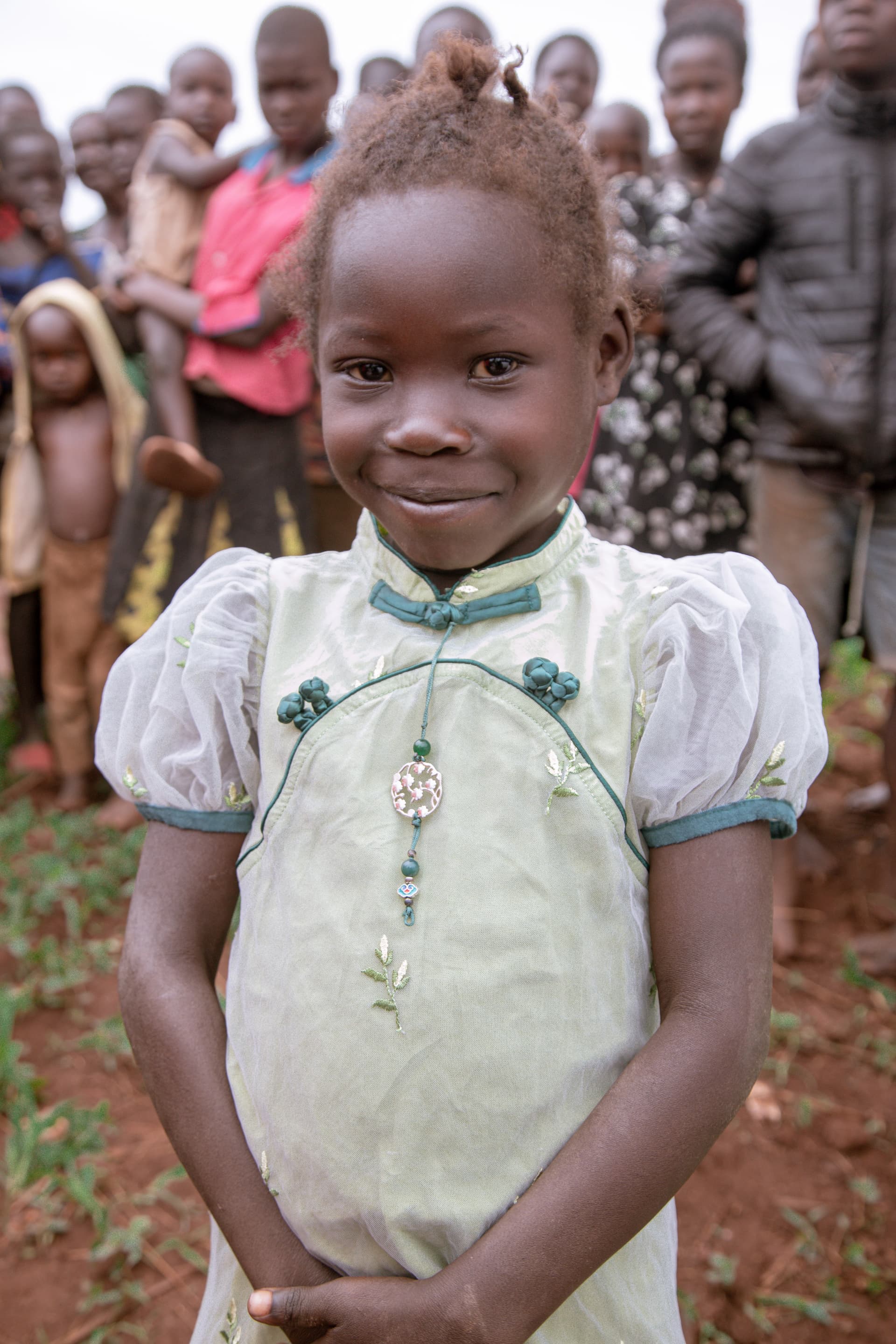 Young girl in a white embroidered dress standing outdoors with a crowd of children behind her.
