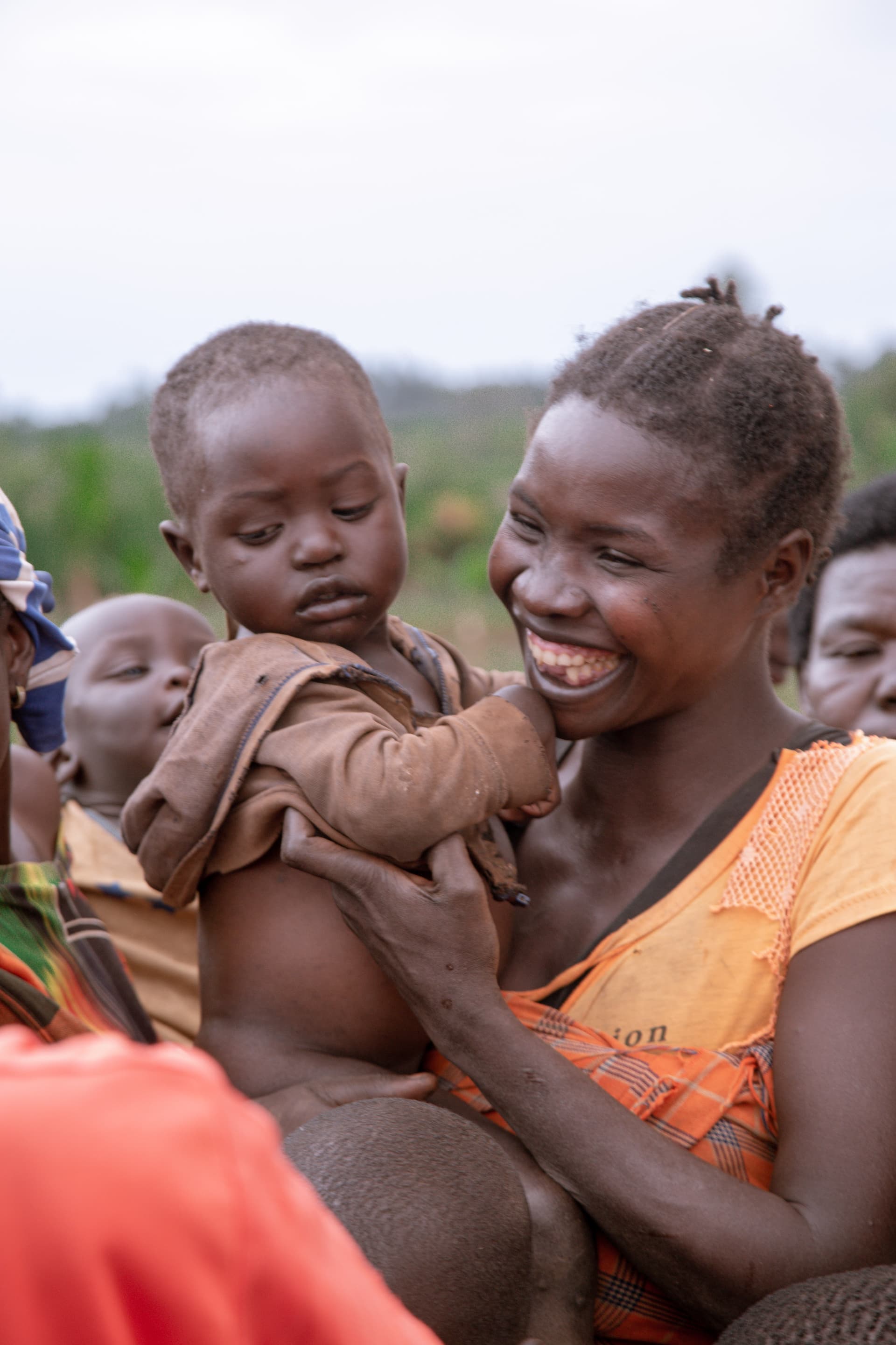 Woman smiling while holding a baby, standing outdoors with others in a village setting.