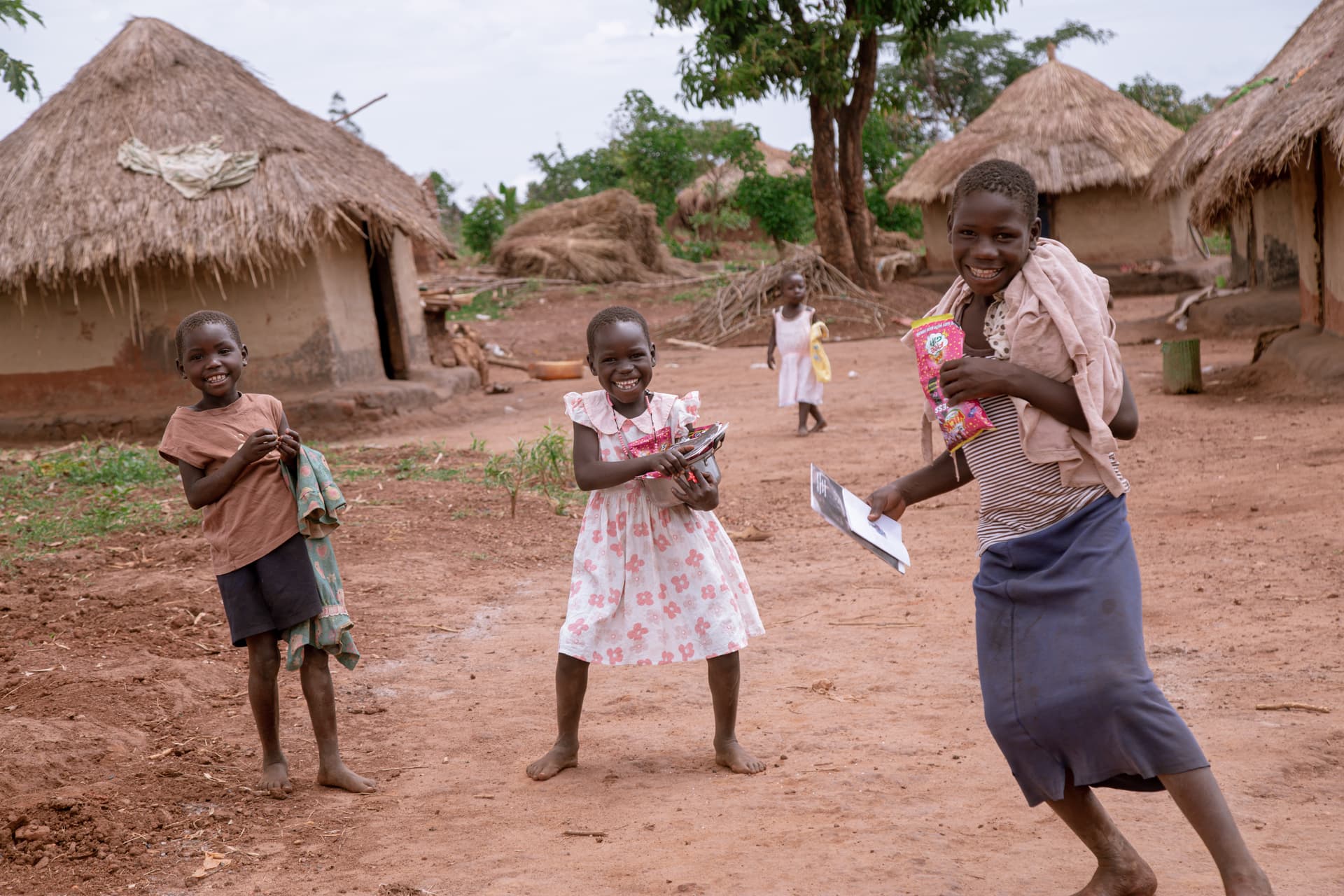 Two girls dance and play joyfully near thatched huts, holding papers in their hands.