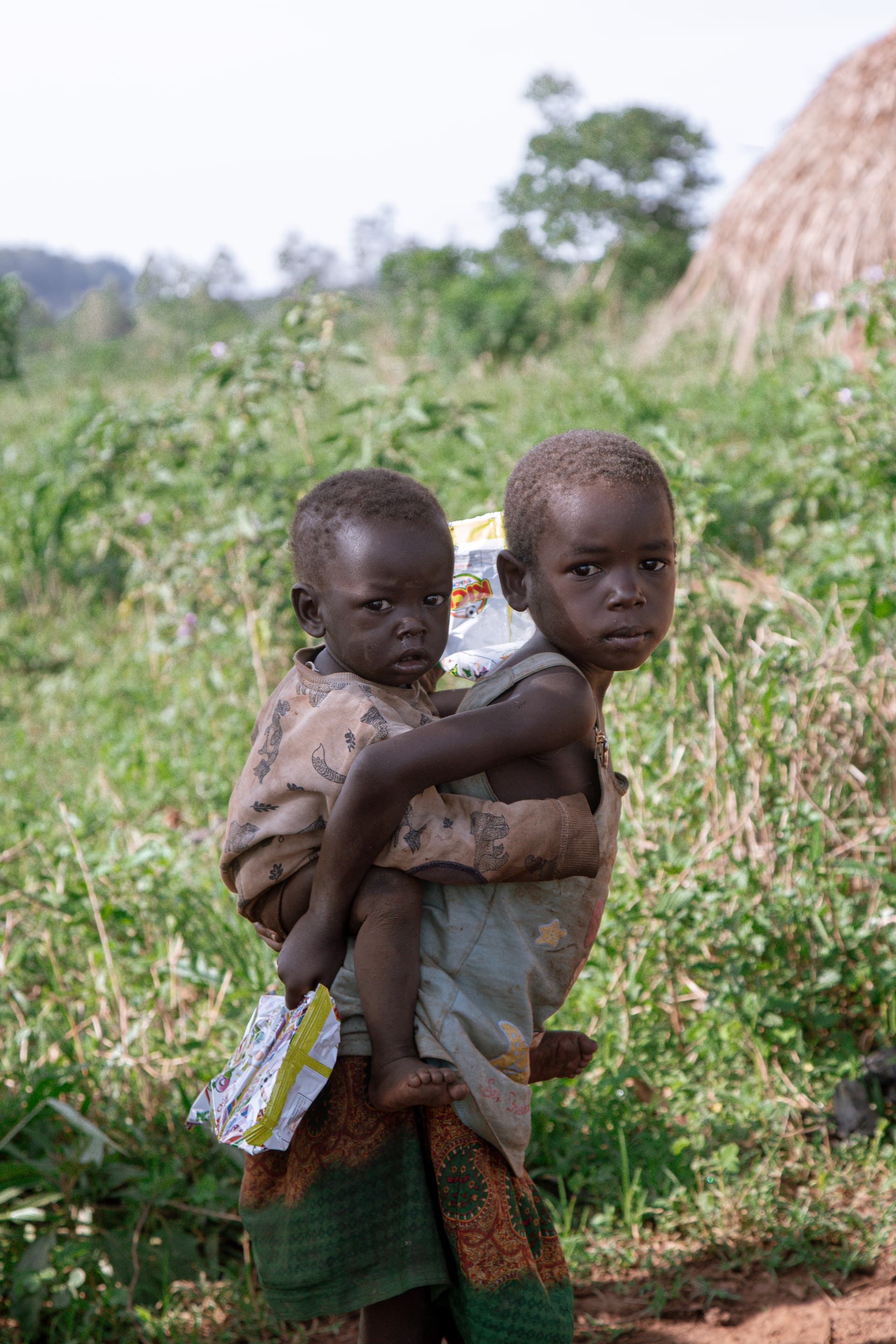 Older child carrying a younger sibling on her back while walking through tall grass near Katulikire village.