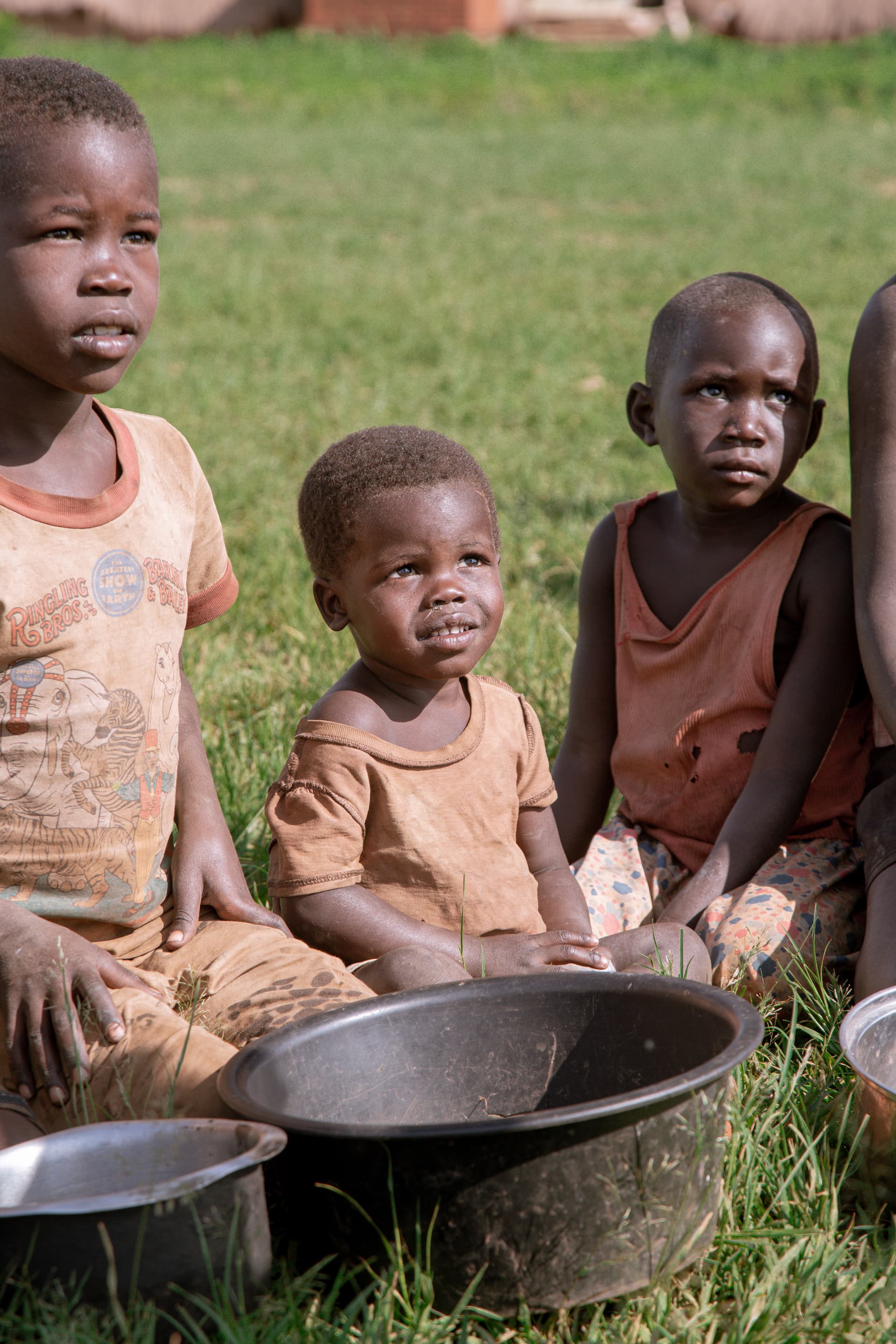 Children sitting on the grass beside empty metal cooking basins, waiting for food distribution.