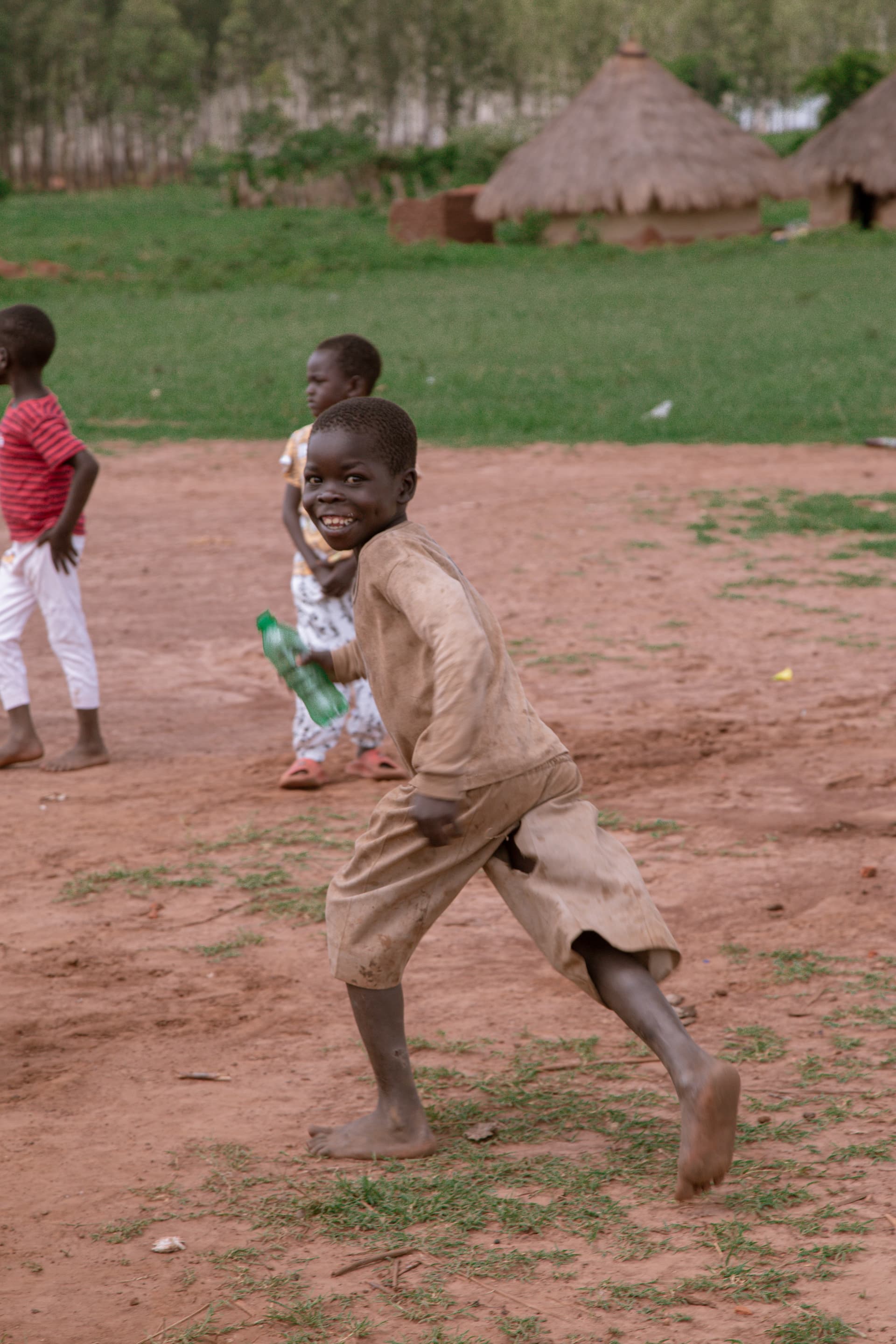 Young boy running across a bare dirt yard with thatched huts in the background.