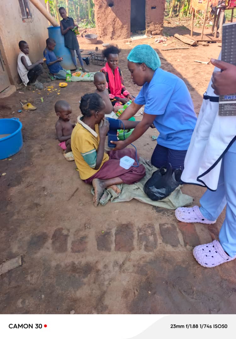 Miracle Medical Center worker in a blue scrub cap examining seated children outdoors near a brick house in Kyampisi Village.