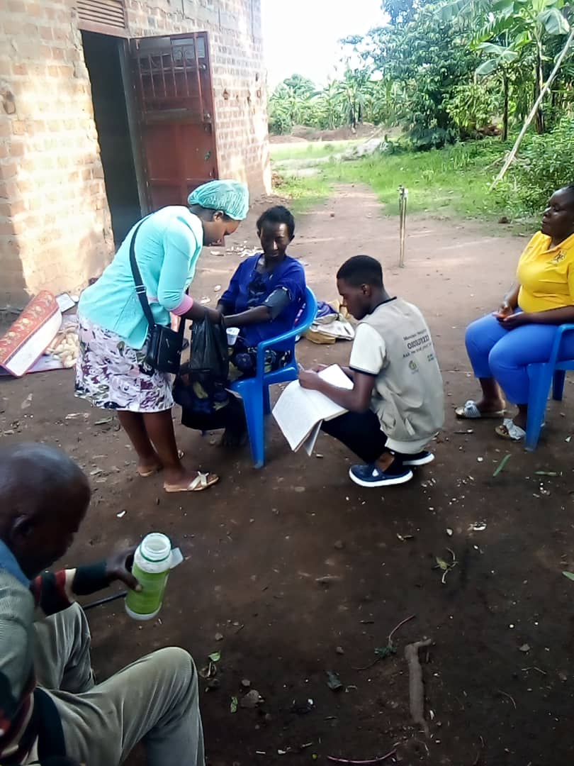 Miracle Medical Center worker in a blue scrub cap examining seated patients outdoors near a brick building.