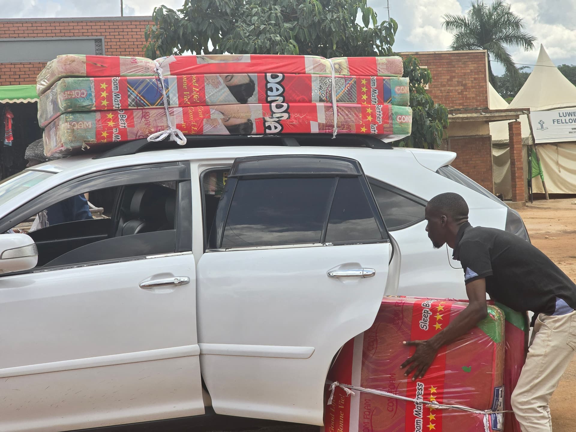 A man loads a large plastic-wrapped mattress bundle into the back seat of a white car.
