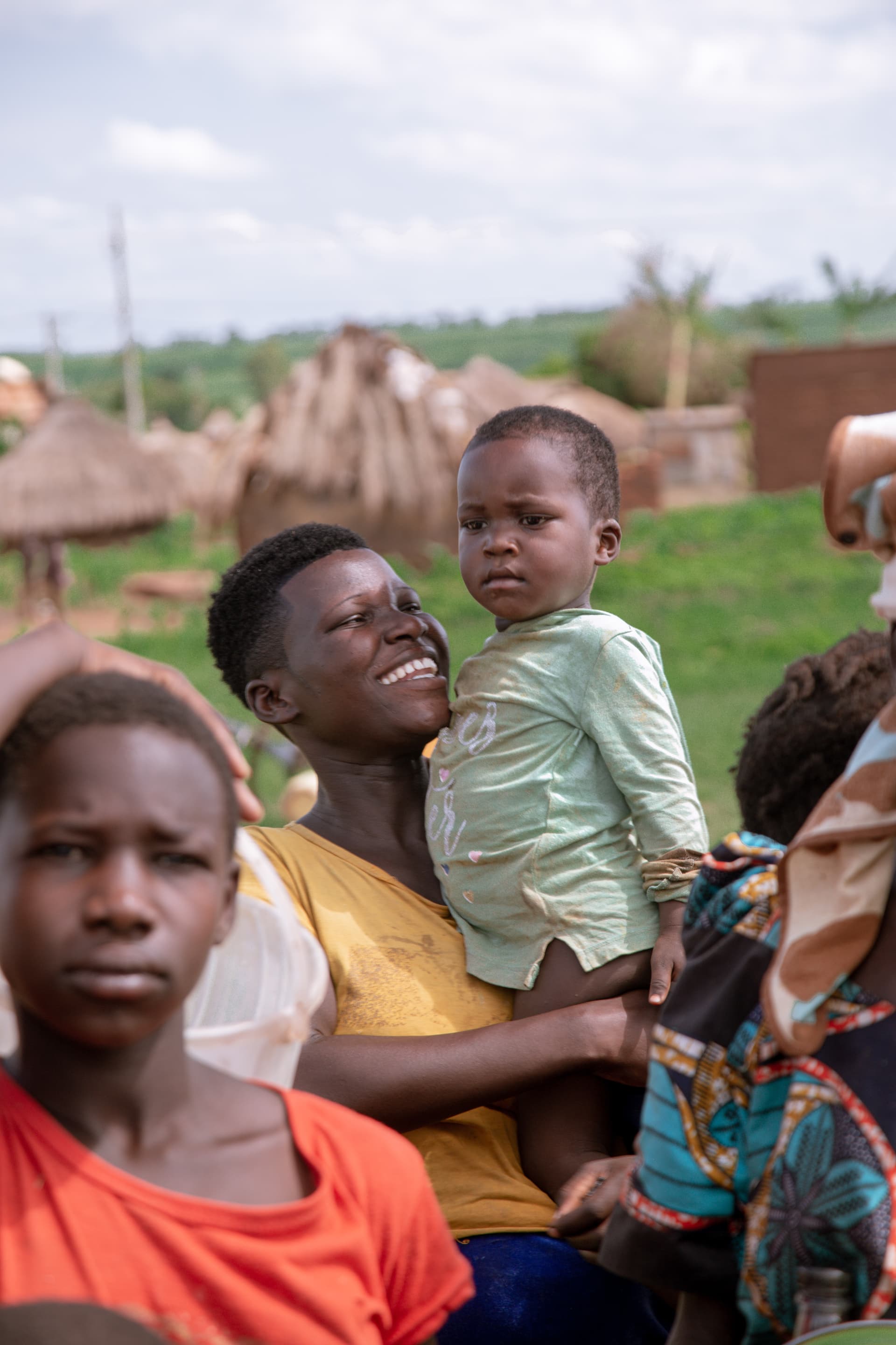 Ugandan mother in an orange top holding a toddler in a mint green outfit, thatched huts behind her.