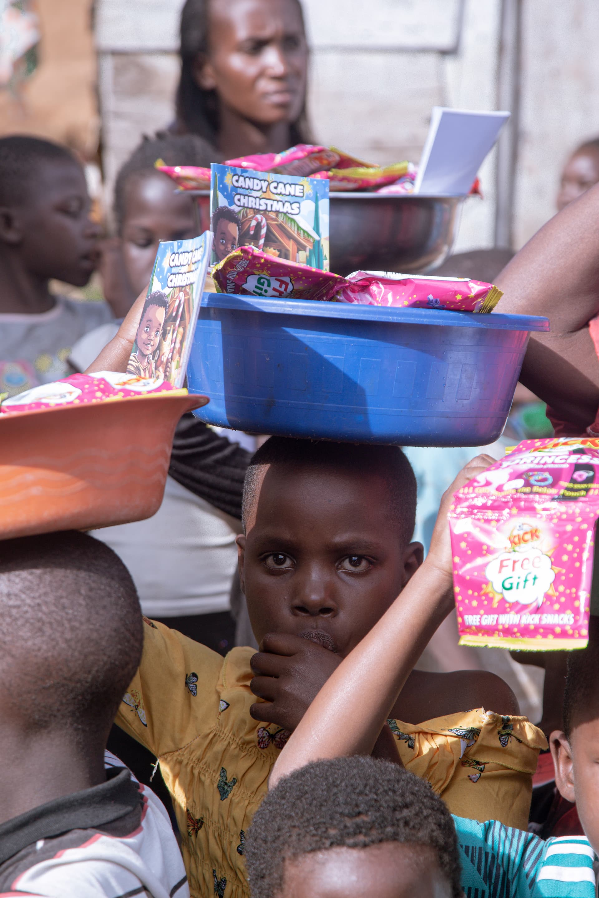 Woman balancing a blue basin loaded with food packages on her head amid a crowd of children.