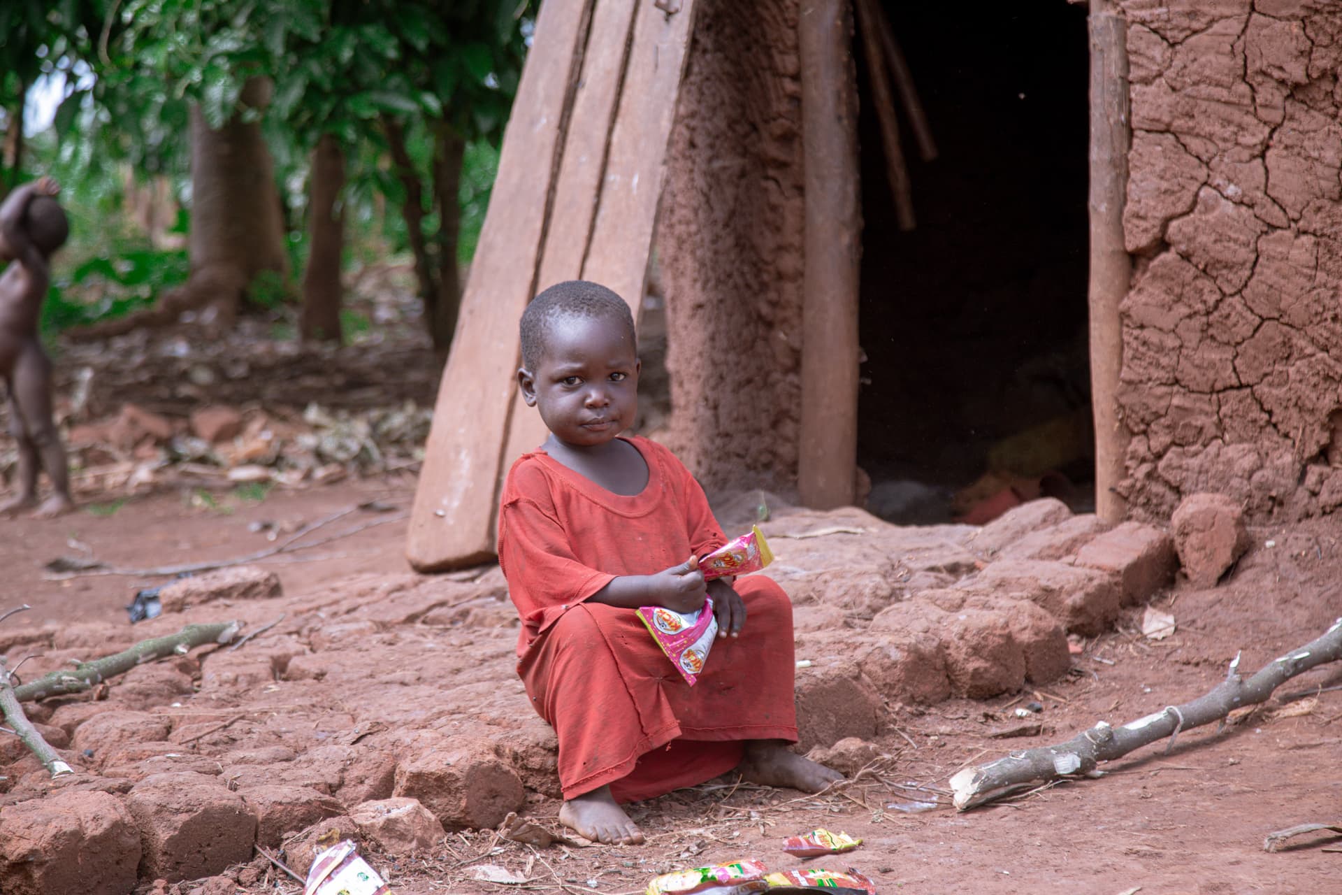 A small child in a red outfit sits alone on the ground near a mud-brick hut.