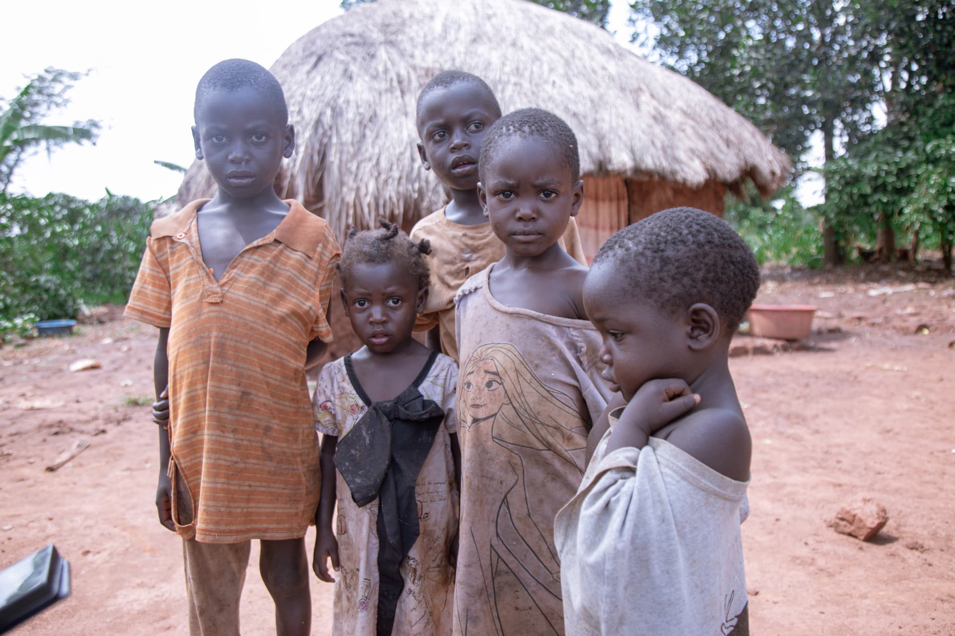 A group of children stands together outdoors in front of a thatched-roof hut.