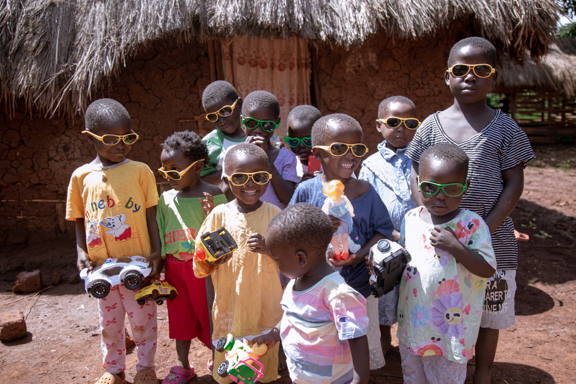 A group of children wearing sunglasses and matching bright patterned outfits stand near a thatched hut.