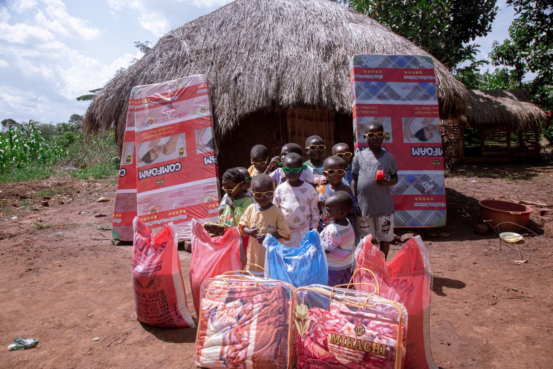 Children gather outside a thatched hut beside red-and-blue wrapped mattress packages delivered by JFM.