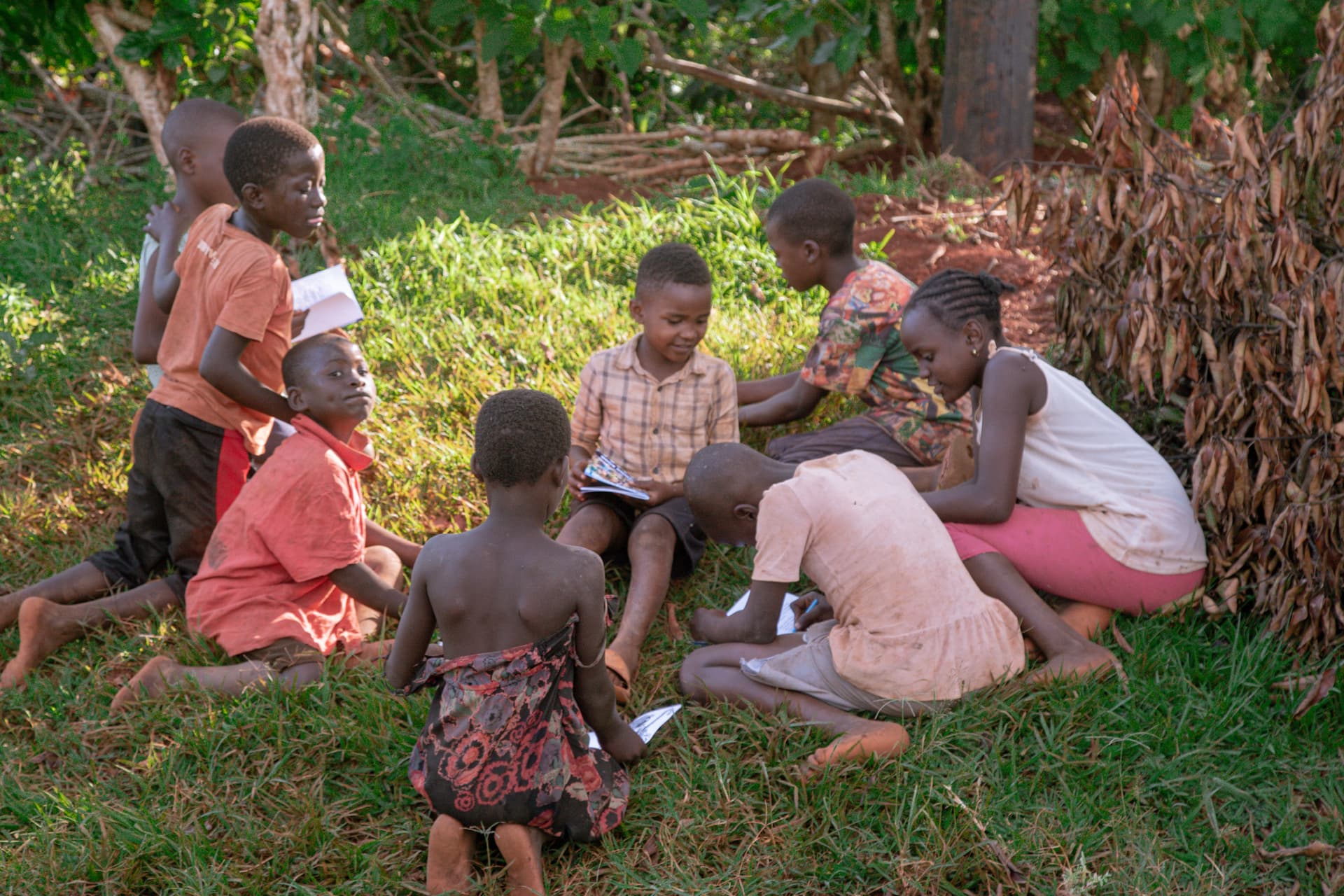 A group of children sits on the grass with an adult, looking together at a coloring book.
