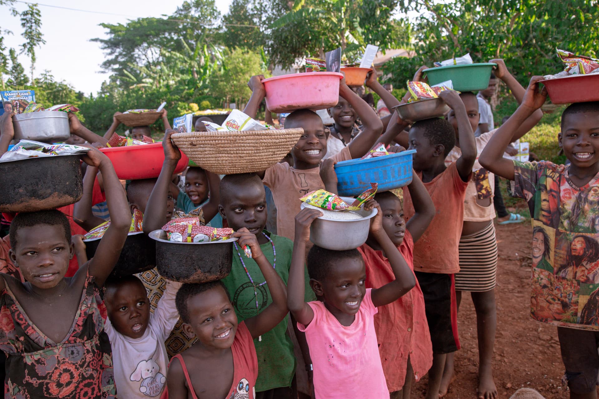 Children carry colorful plastic basins and bowls of food balanced on their heads outdoors.