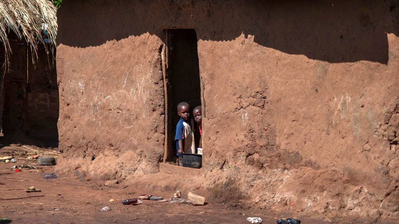 A small child stands alone in the doorway of a mud-brick home with a thatched roof.