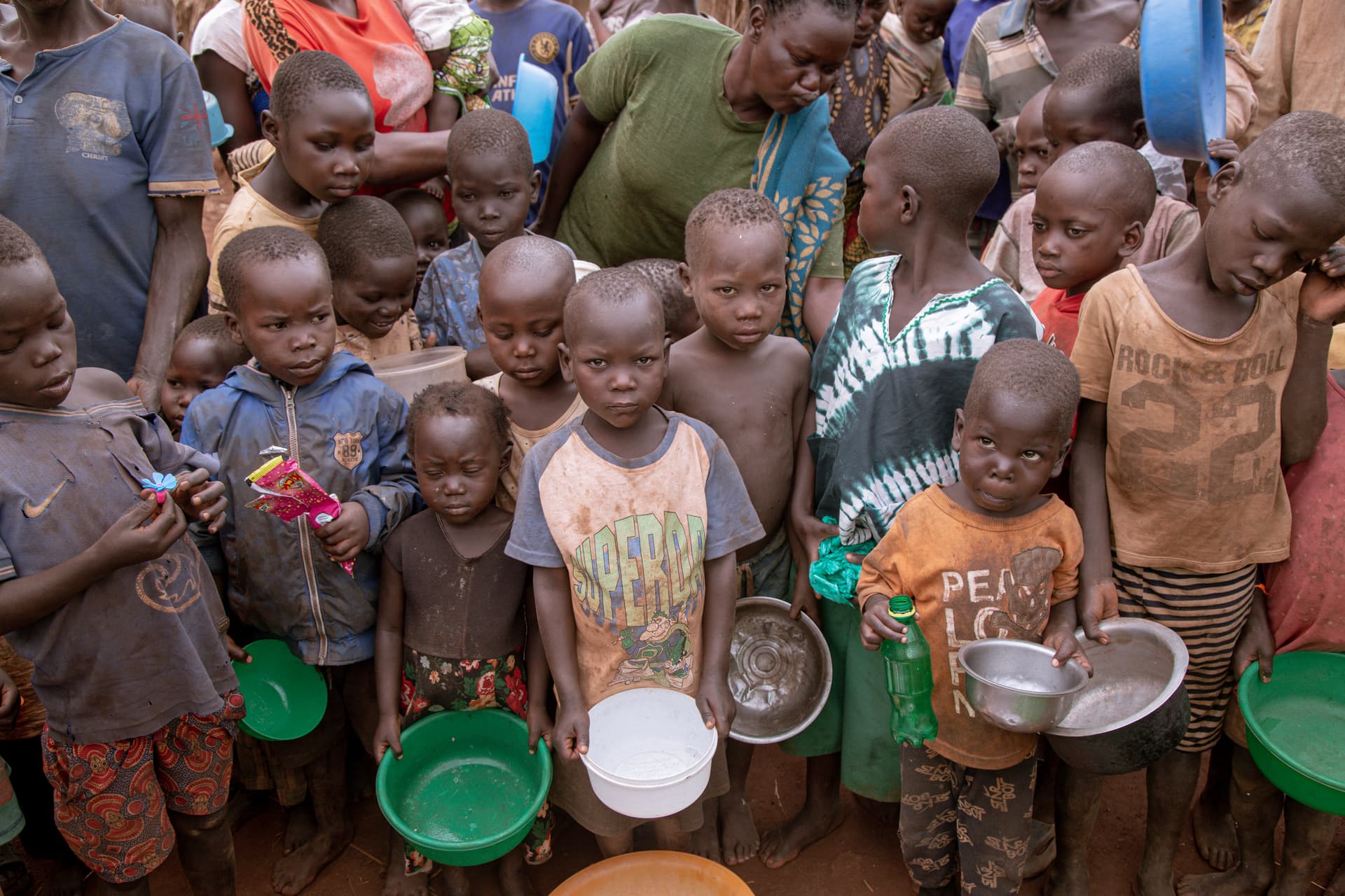Children stand in a line holding metal bowls and plastic cups, waiting at a JFM food distribution.