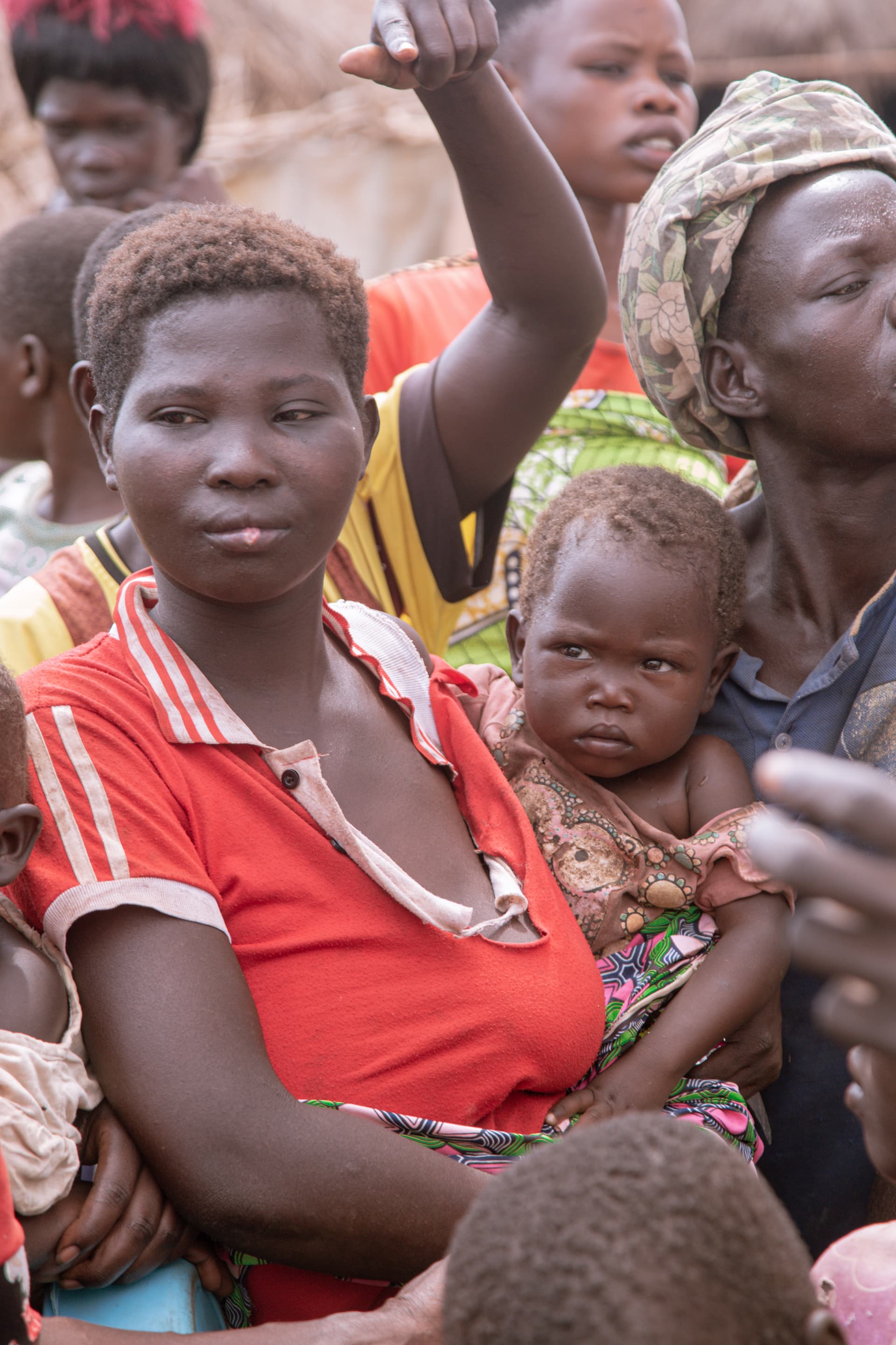 A mother in a red shirt holds a baby while standing in a group of women and children.