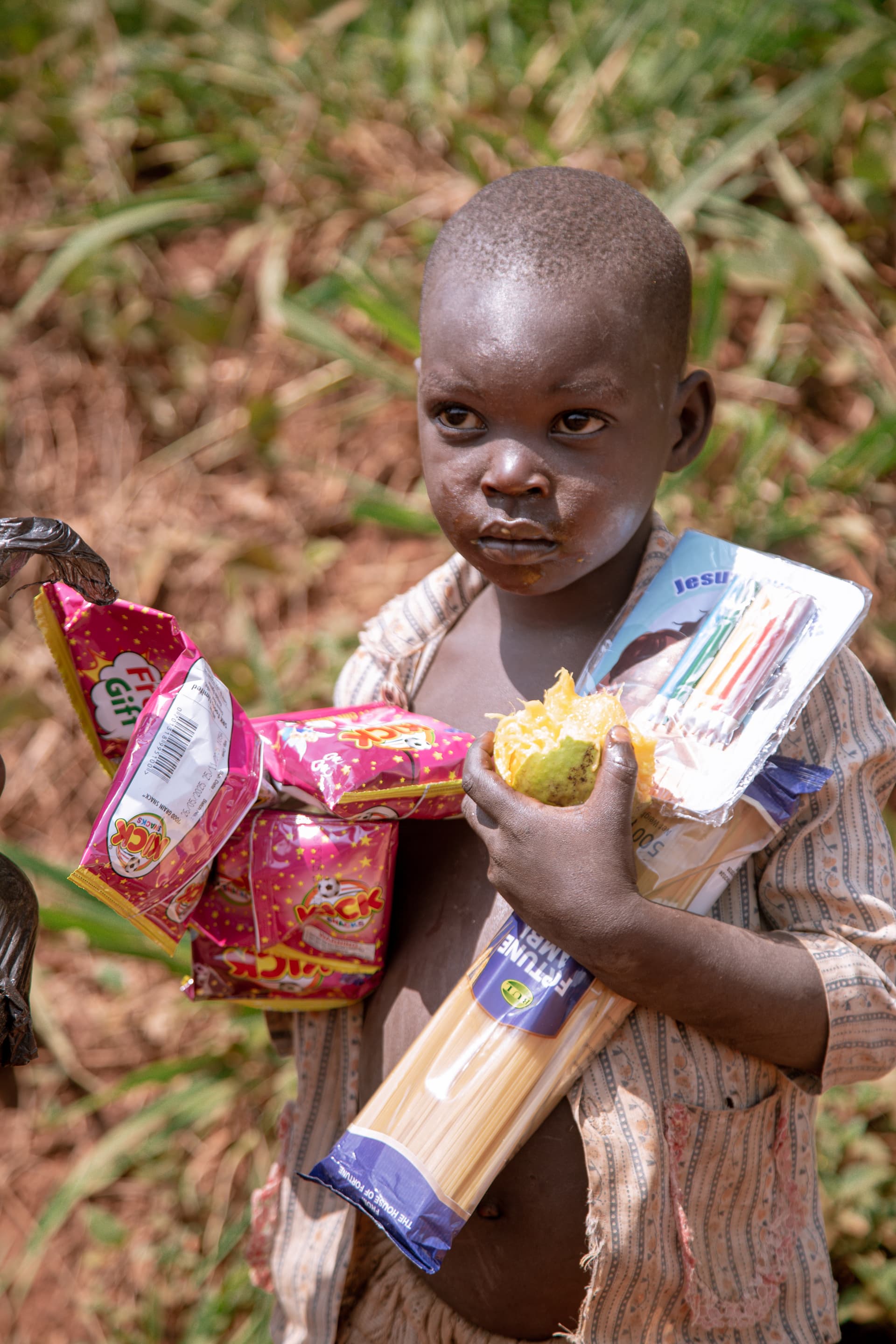 Toddler holding pink and yellow food packets, standing outdoors in the grass.