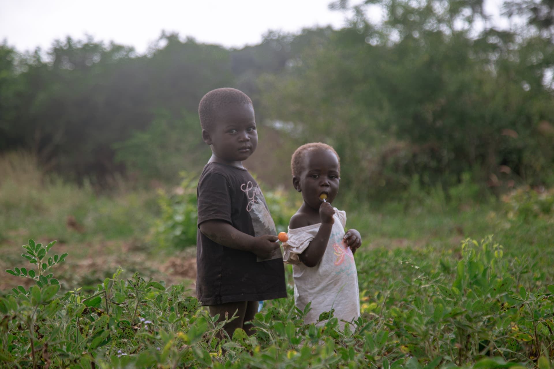 Two small children, James Oyot's, standing together outdoors in a hazy grassy field.