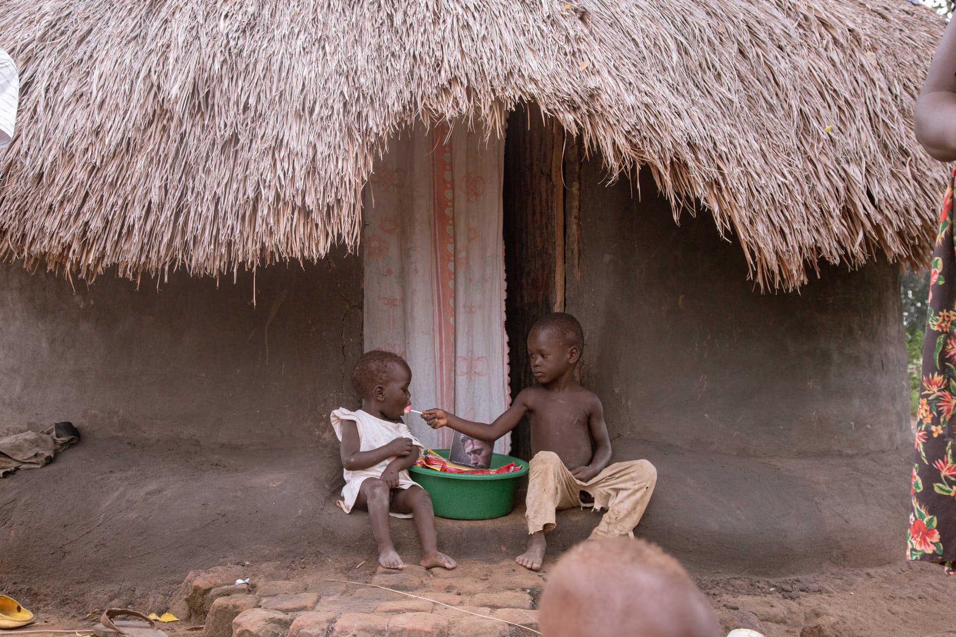 Two children sitting outside a thatched hut near a green basin of food packets.