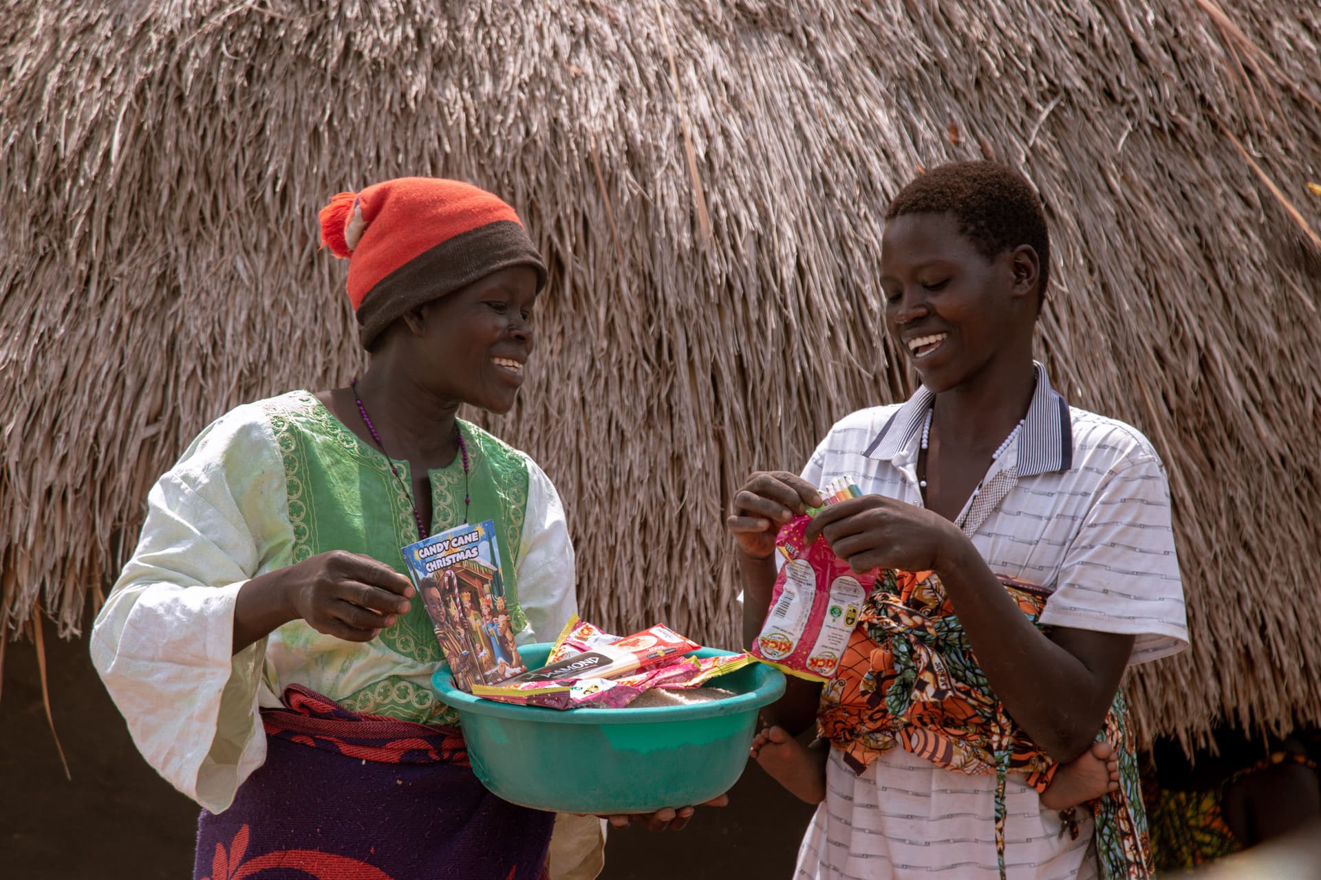 Two women, one older, standing together under a thatched roof holding basins of food items.