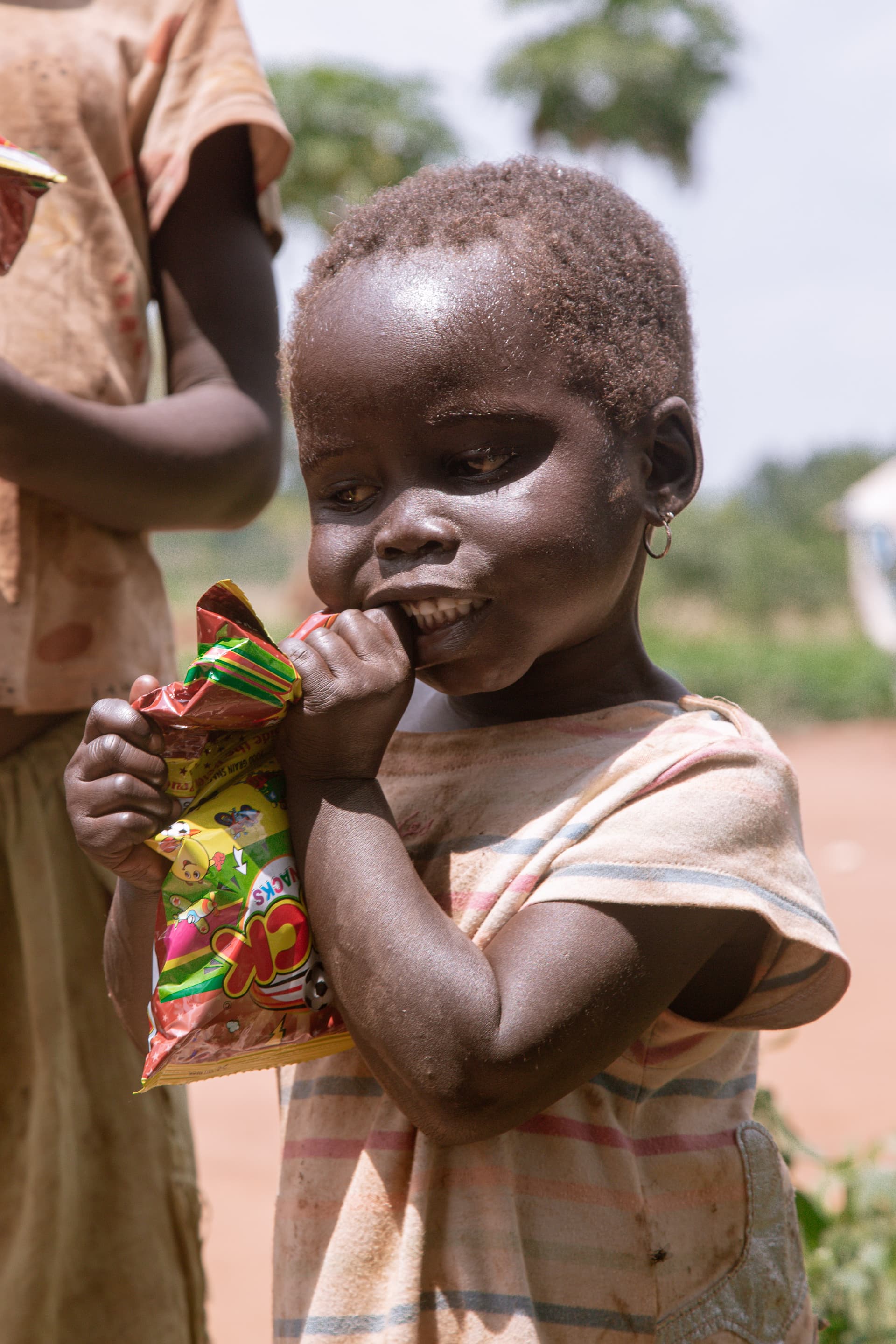 An adult holds a toddler who eats a food snack outdoors.