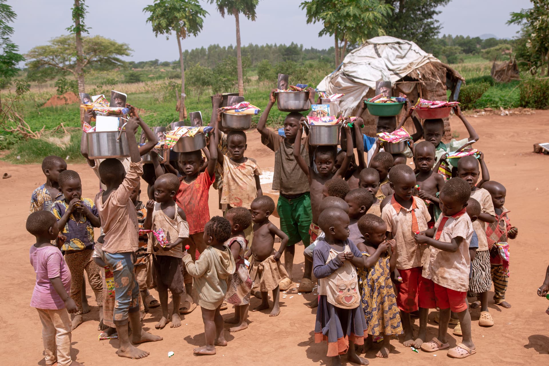 Line of children walking together through the village, several balancing food containers on their heads.