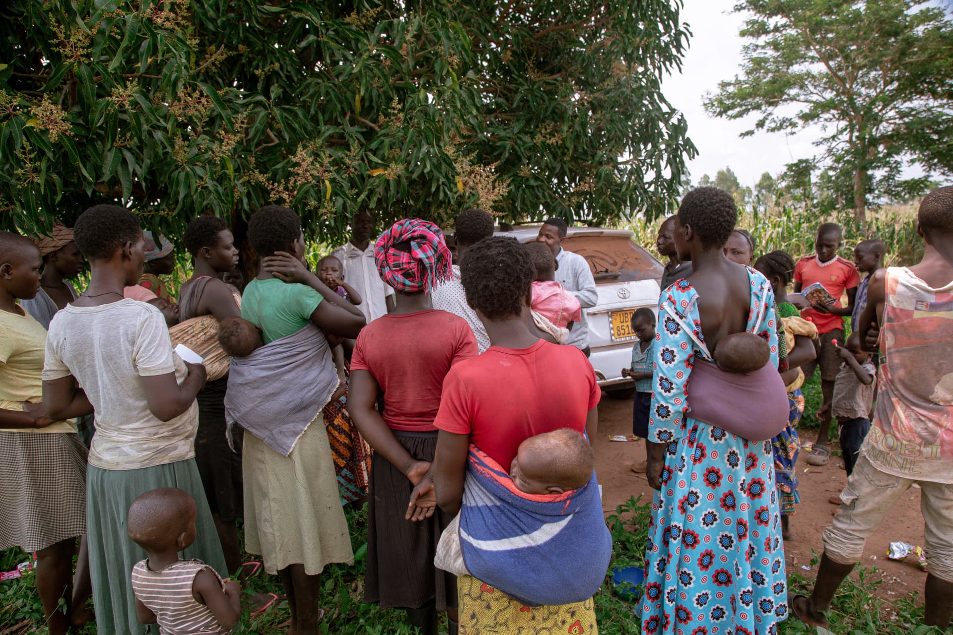 Group of women and children standing in line outdoors, some holding babies, waiting during an outreach in Ogwal.