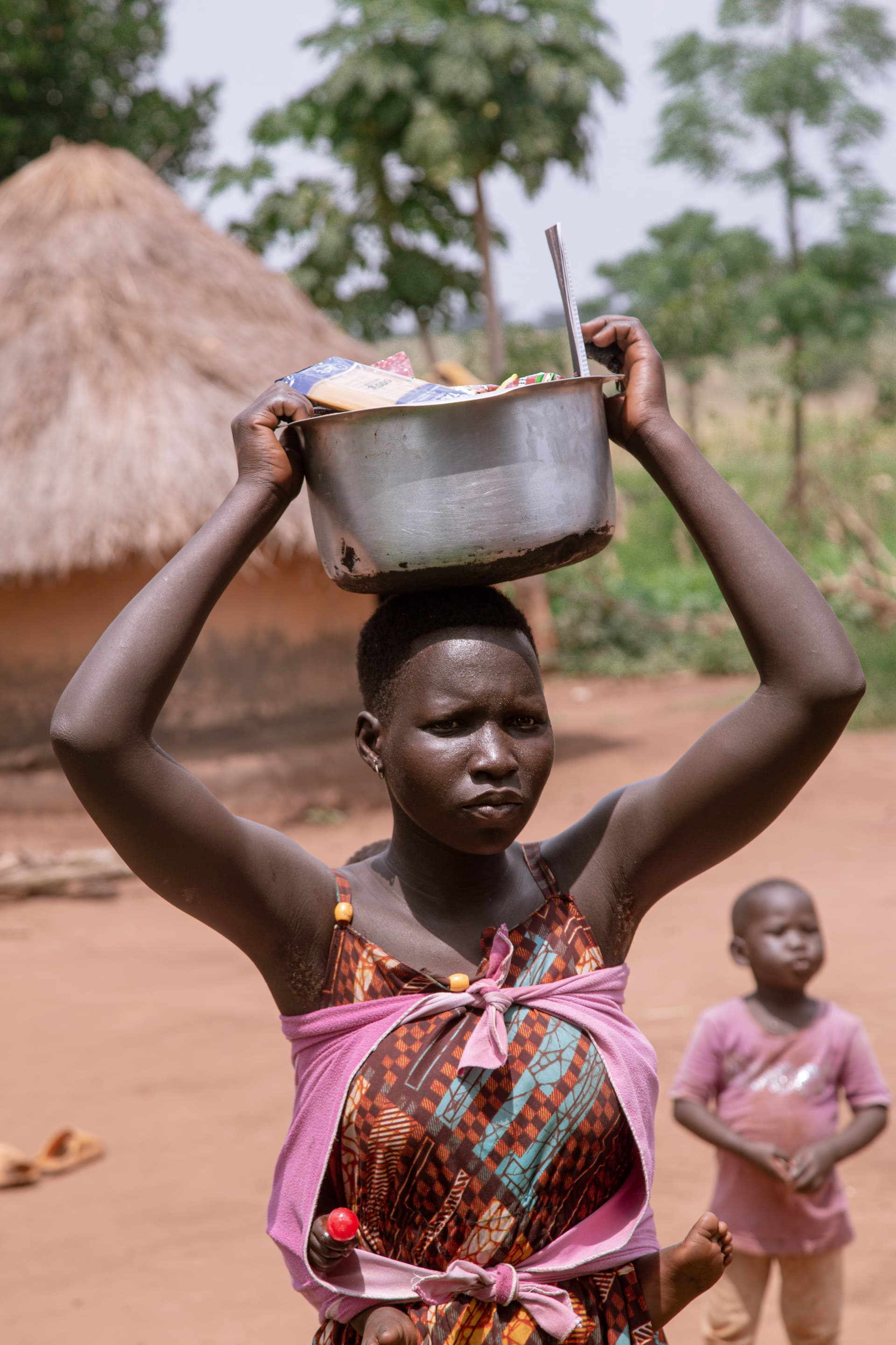 Woman carrying a metal pot of food items on her head with a young girl in a pink dress walking behind her.