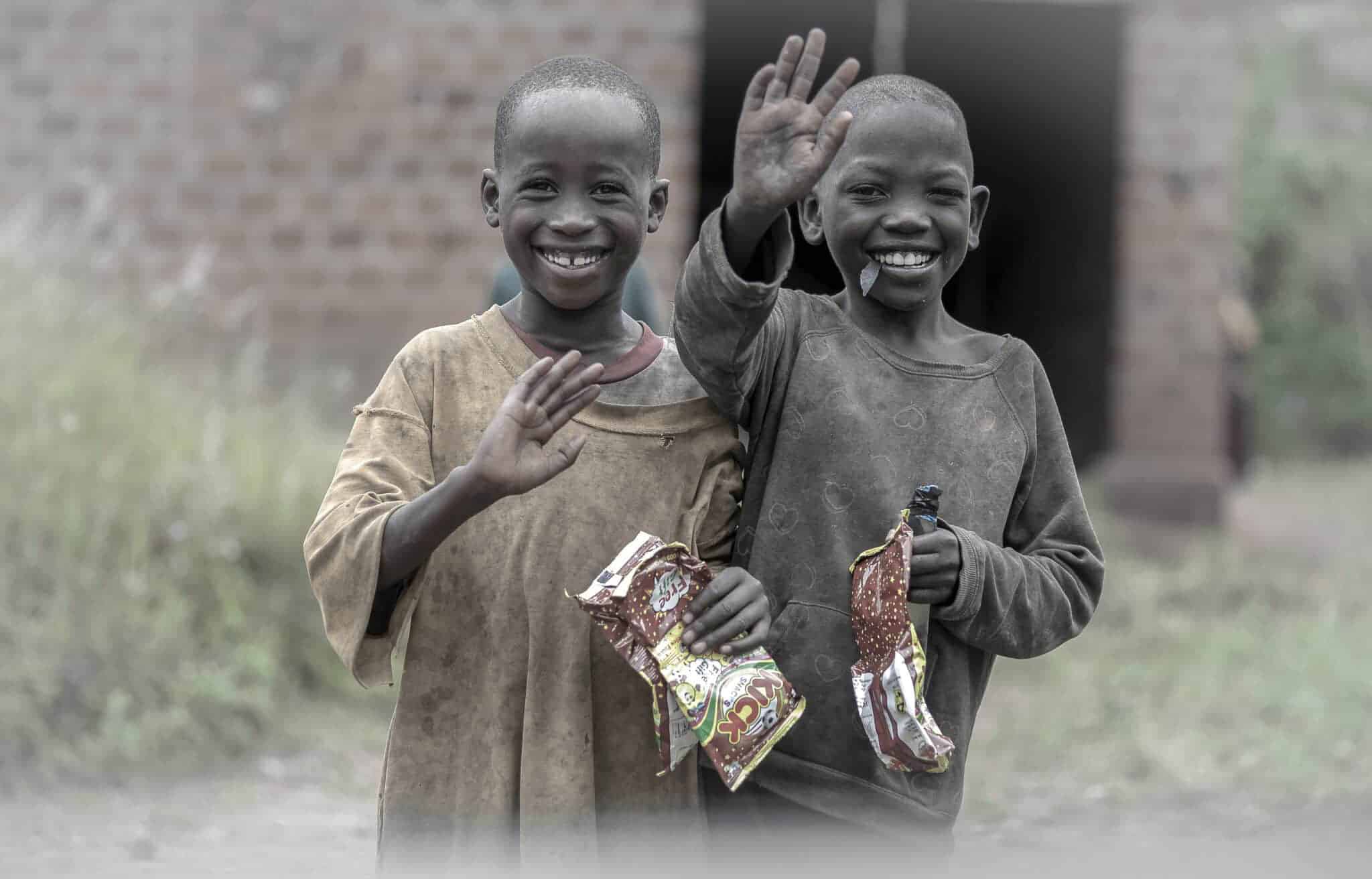 Black-and-white photo of two boys waving and smiling outside a building while holding food packets.
