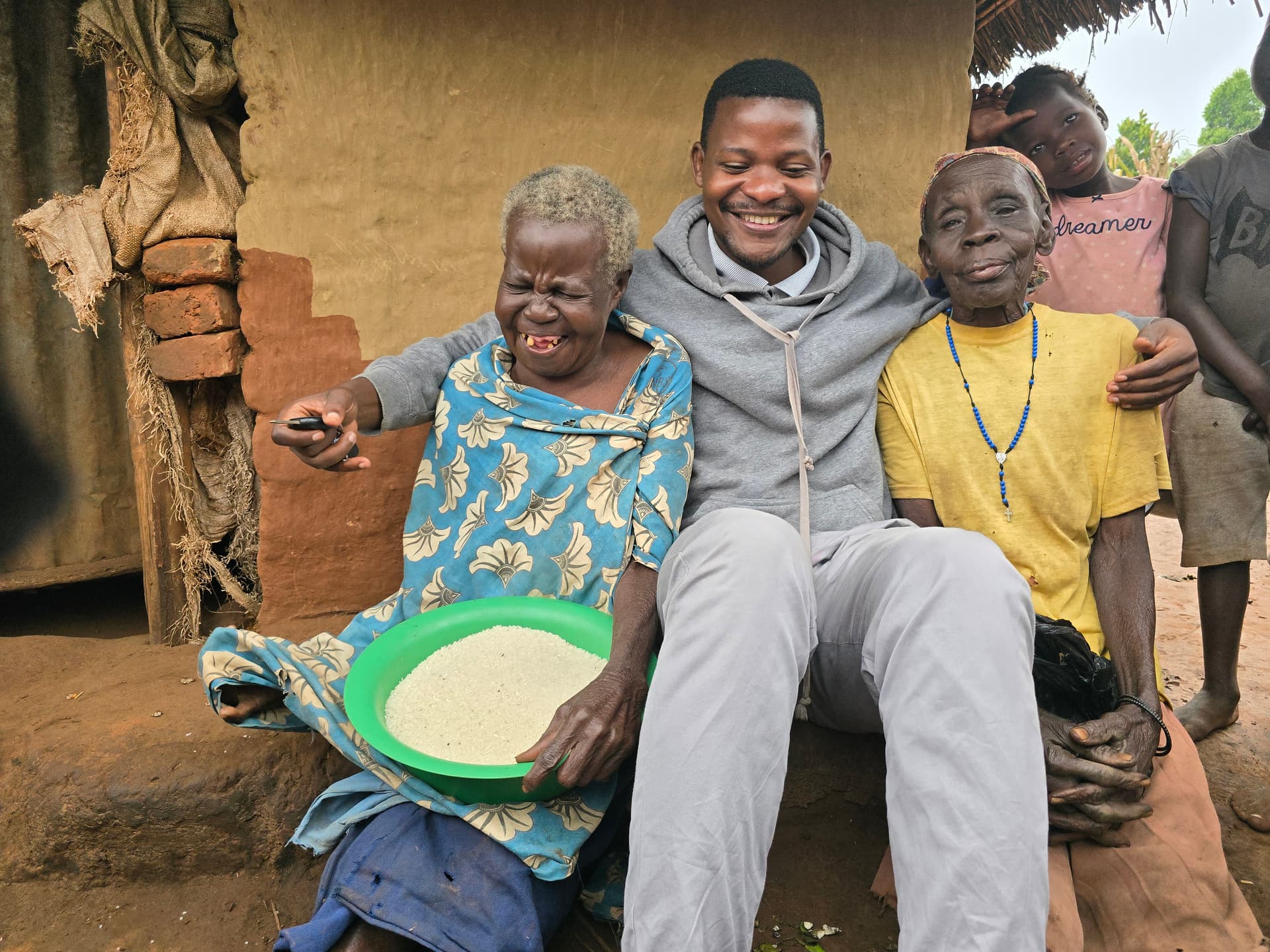 A man sits with two elderly women, one holding a green bowl, in front of a mud house.