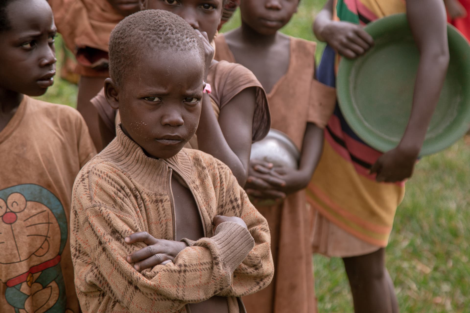 A group of children stand together outdoors near Taalari, one boy holding food items and a green bowl.