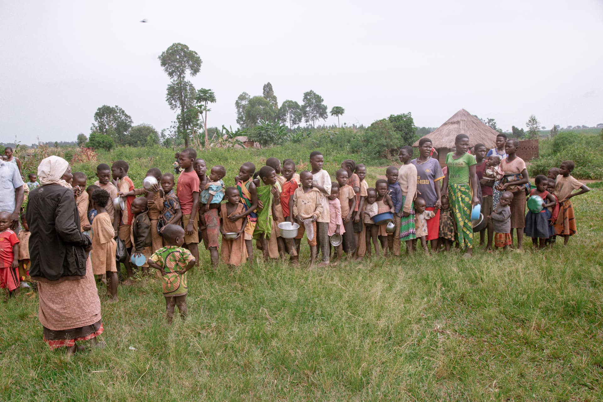 A line of Ugandan women and children stand outdoors waiting, one carrying a bundle on her head.