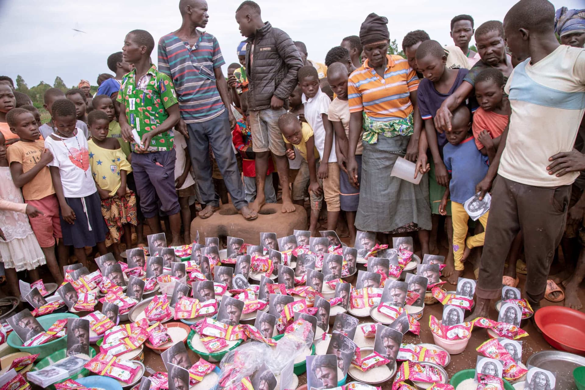 A crowd of Ugandan children and adults stand behind rows of packaged food gifts laid on the ground.