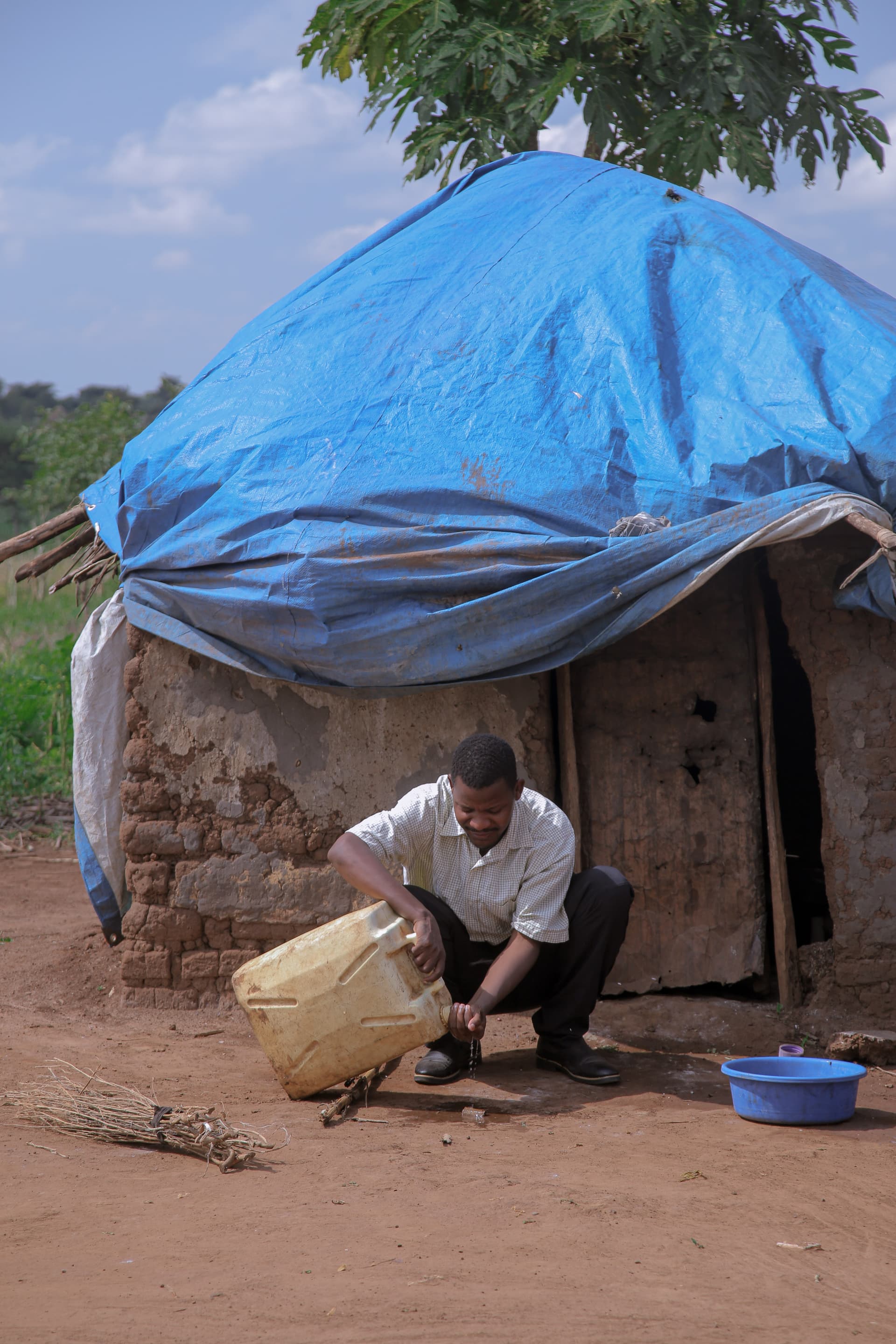 A man crouches to fill a jerry can with water outside a mud-brick hut with a blue tarp roof.