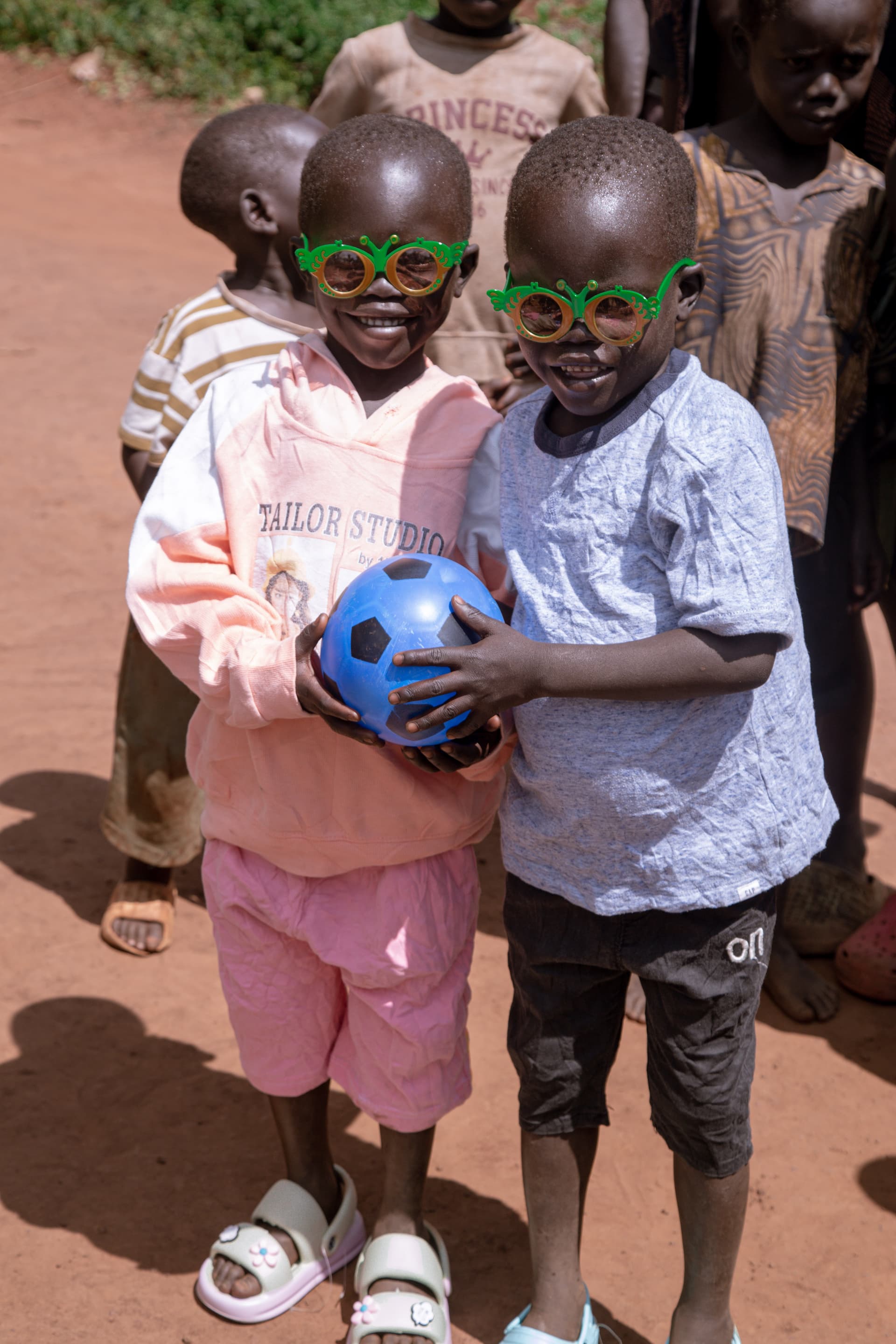Two boys wear new colorful clothes and green sunglasses, one holding a blue soccer ball, among other children.