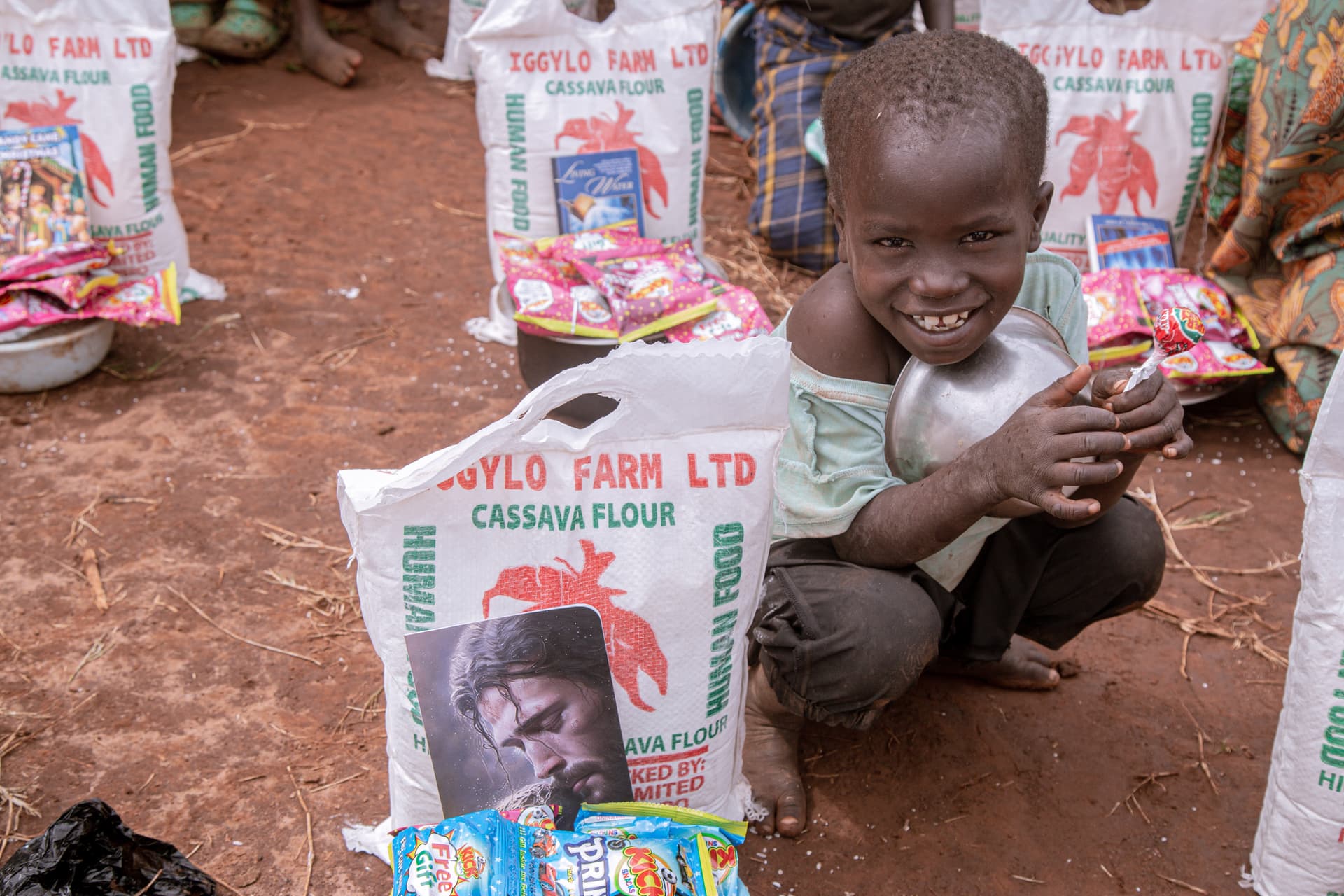 A boy kneels smiling behind a sack of cassava flour holding a printed photo card.