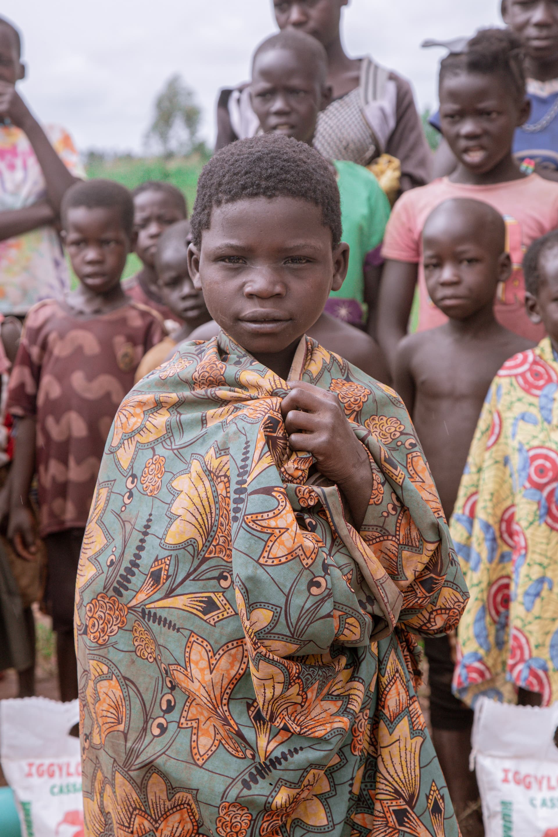 A child wrapped in a colorful blanket stands amid a crowd gathered outdoors.