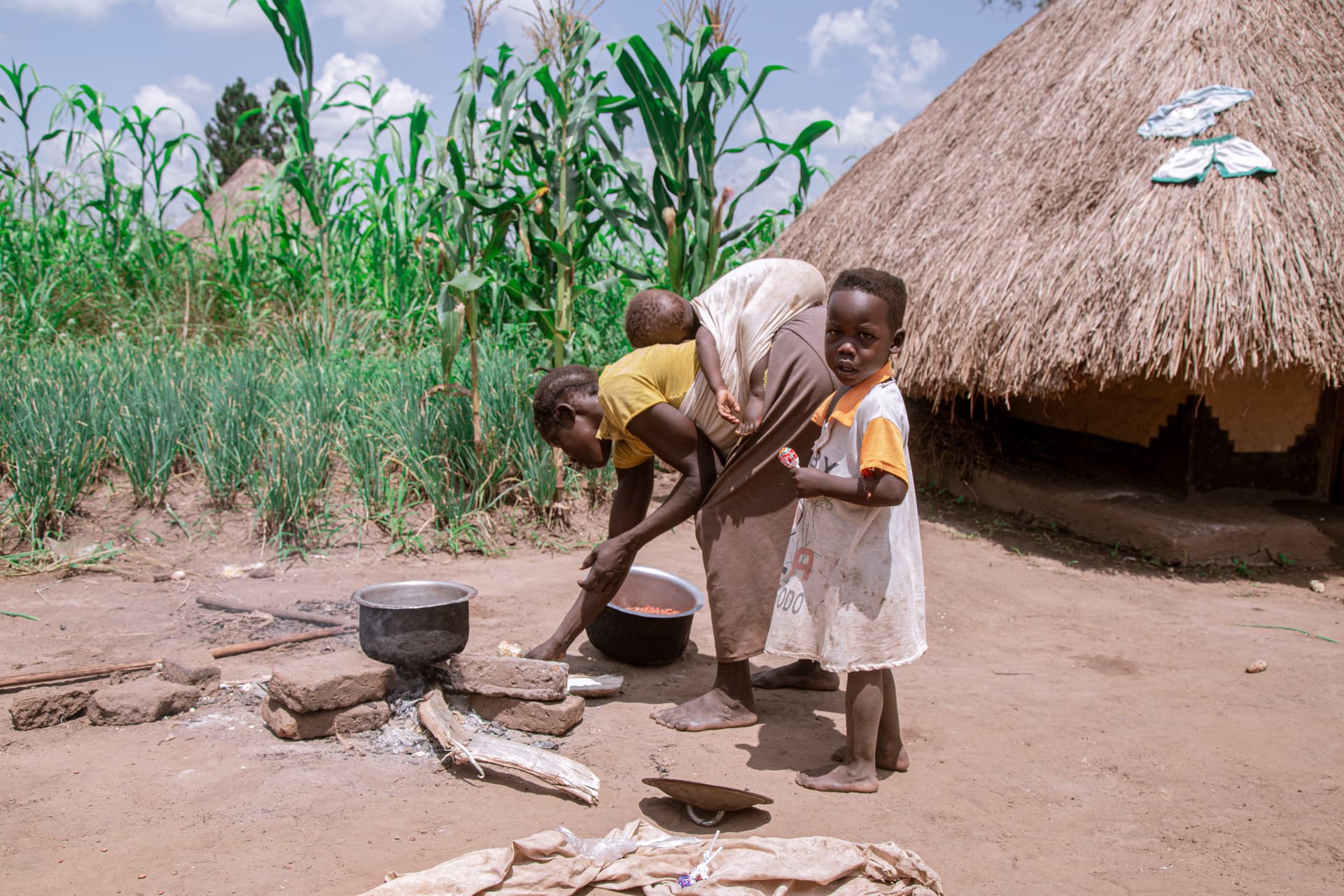 A woman and children cook beans over an open fire outside a thatched hut near cornstalks.