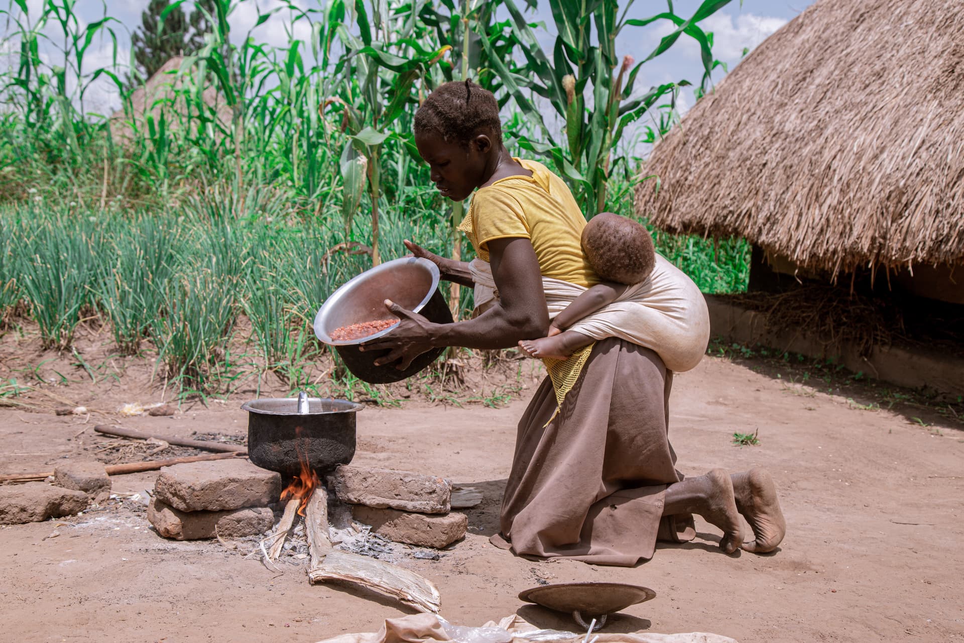 A woman kneeling to cook over an open fire with a baby tied on her back, thatched hut behind.
