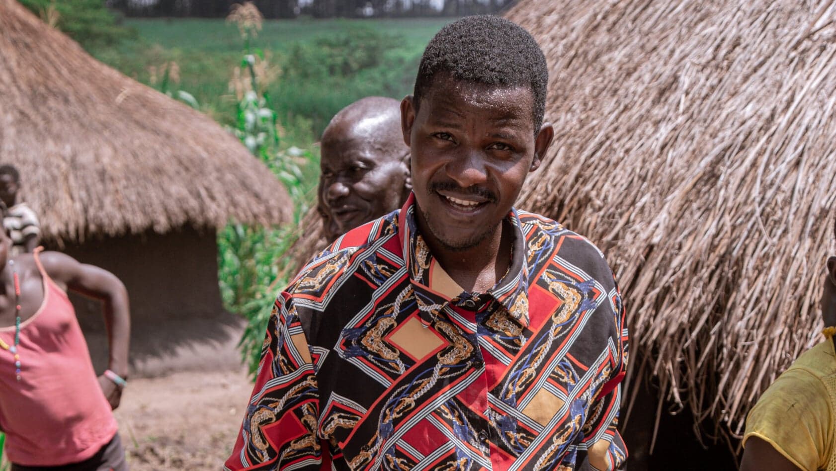 A man in a colorful patterned shirt smiles near thatched-roof huts in a Ugandan village.