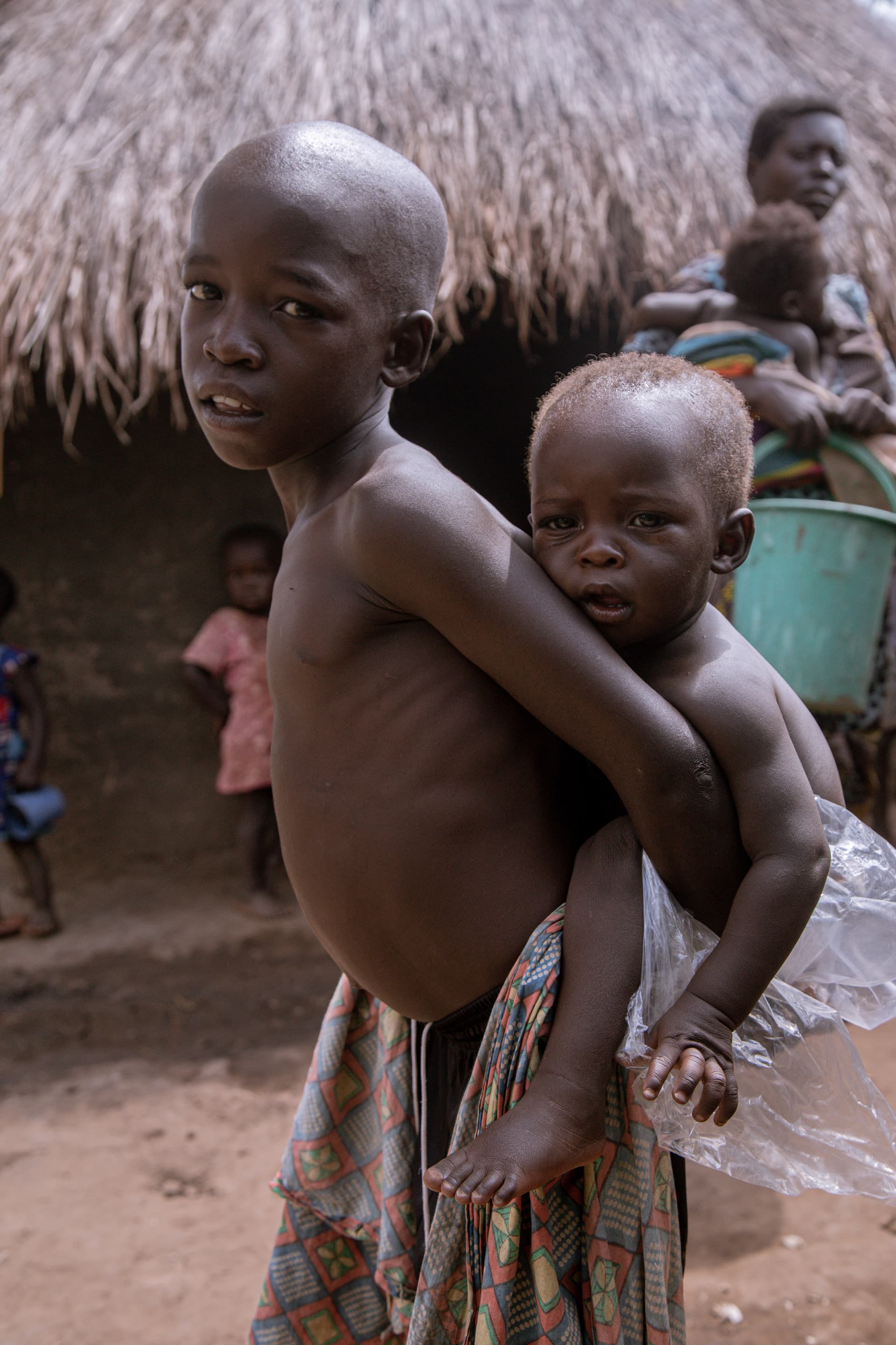A thin child carries a baby on her back beside a thatched hut in Zombo village.