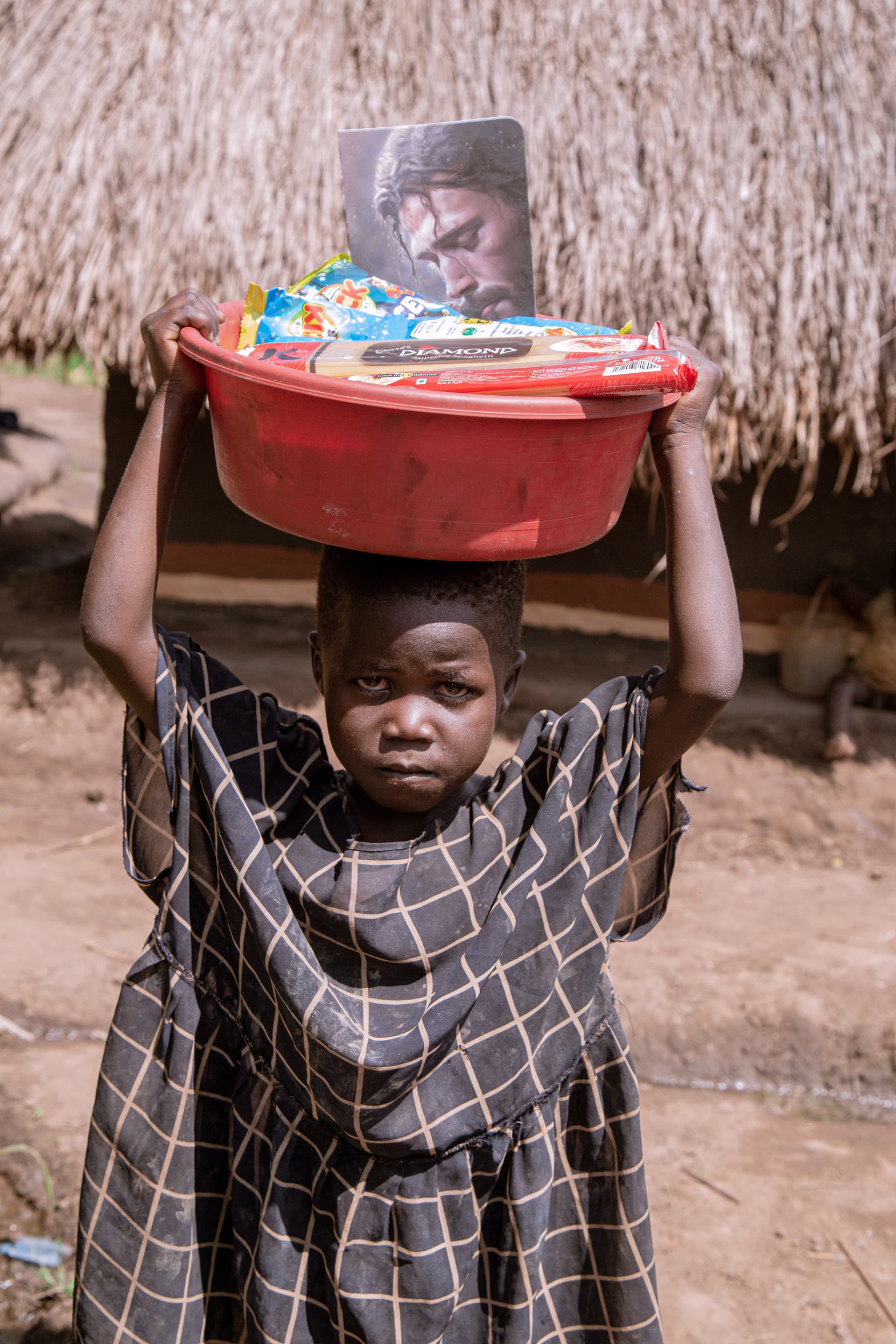A girl carries a red basin with a Jesus Loves Me tract on her head, standing near a thatched roof structure.