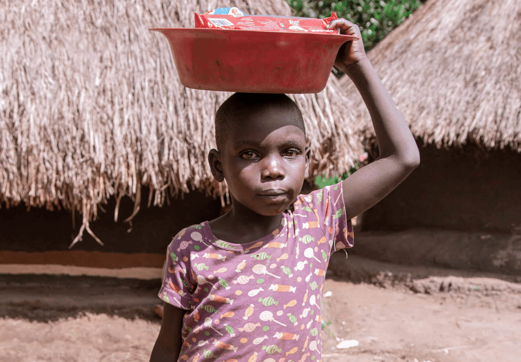 A girl in a pink dress carries a red basin of food on her head near a thatched hut.