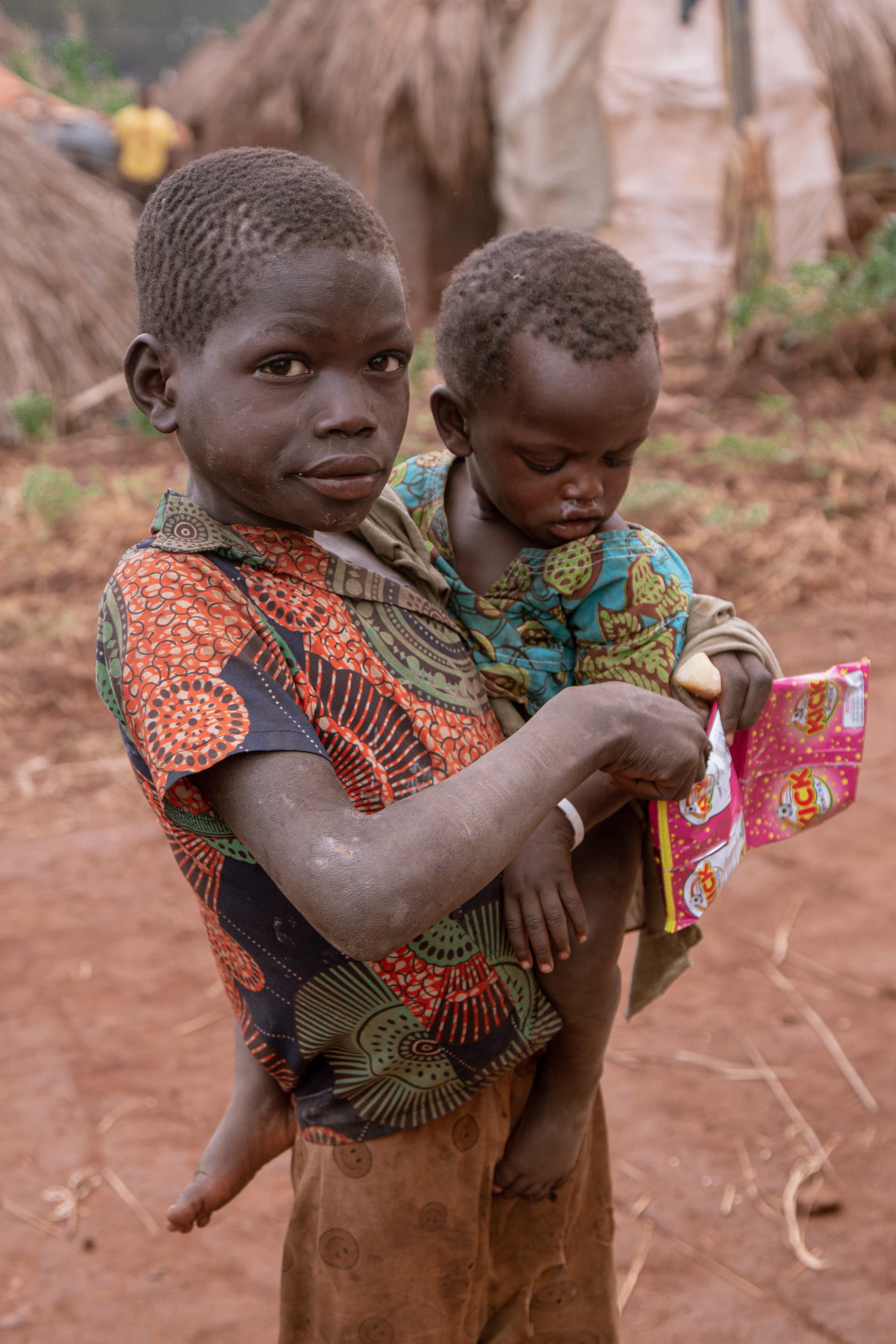 An older child carries a younger sibling on her back while holding a pink JFM food package.