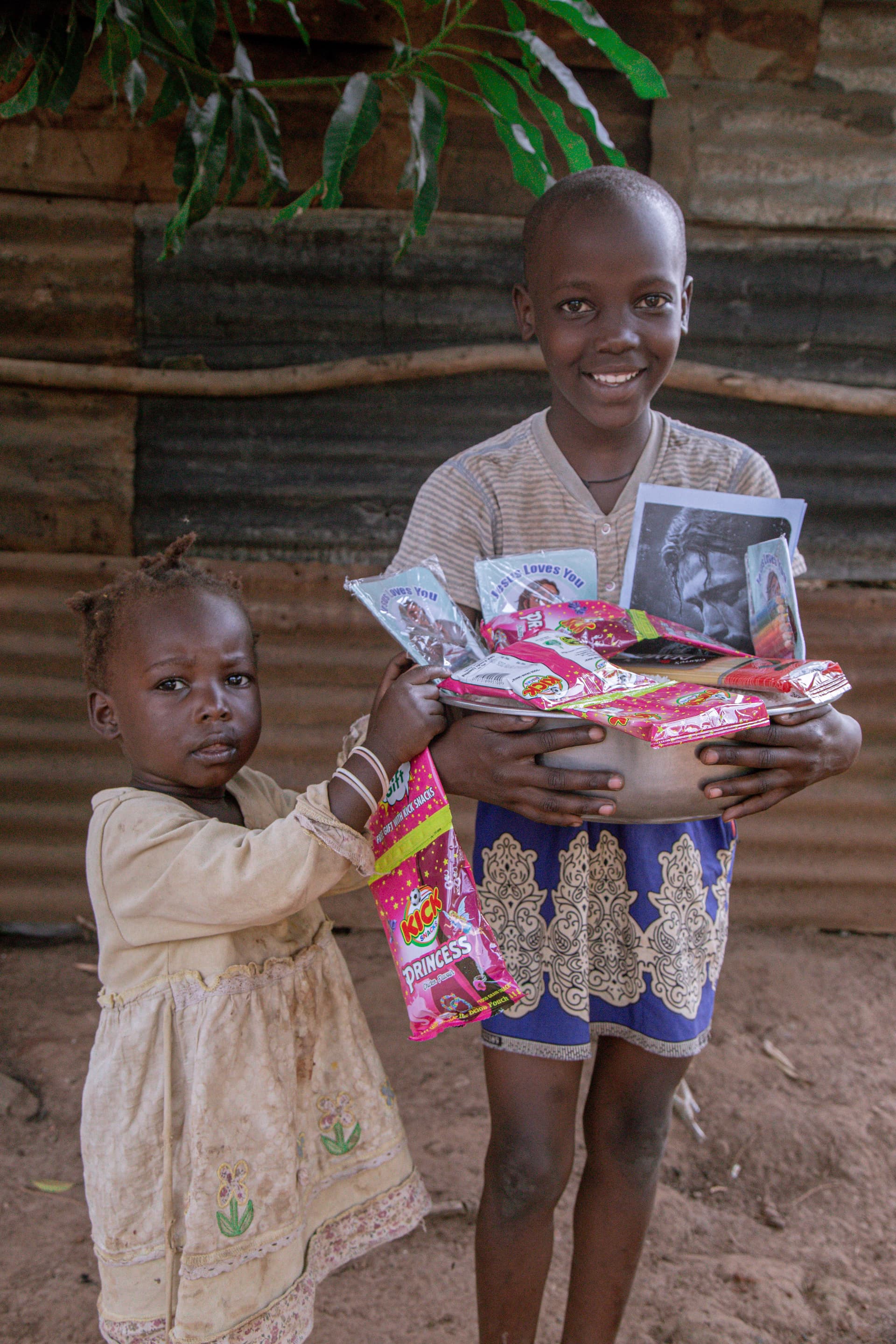 An older boy and a young girl hold a pink food package and photo card under a thatched shelter.