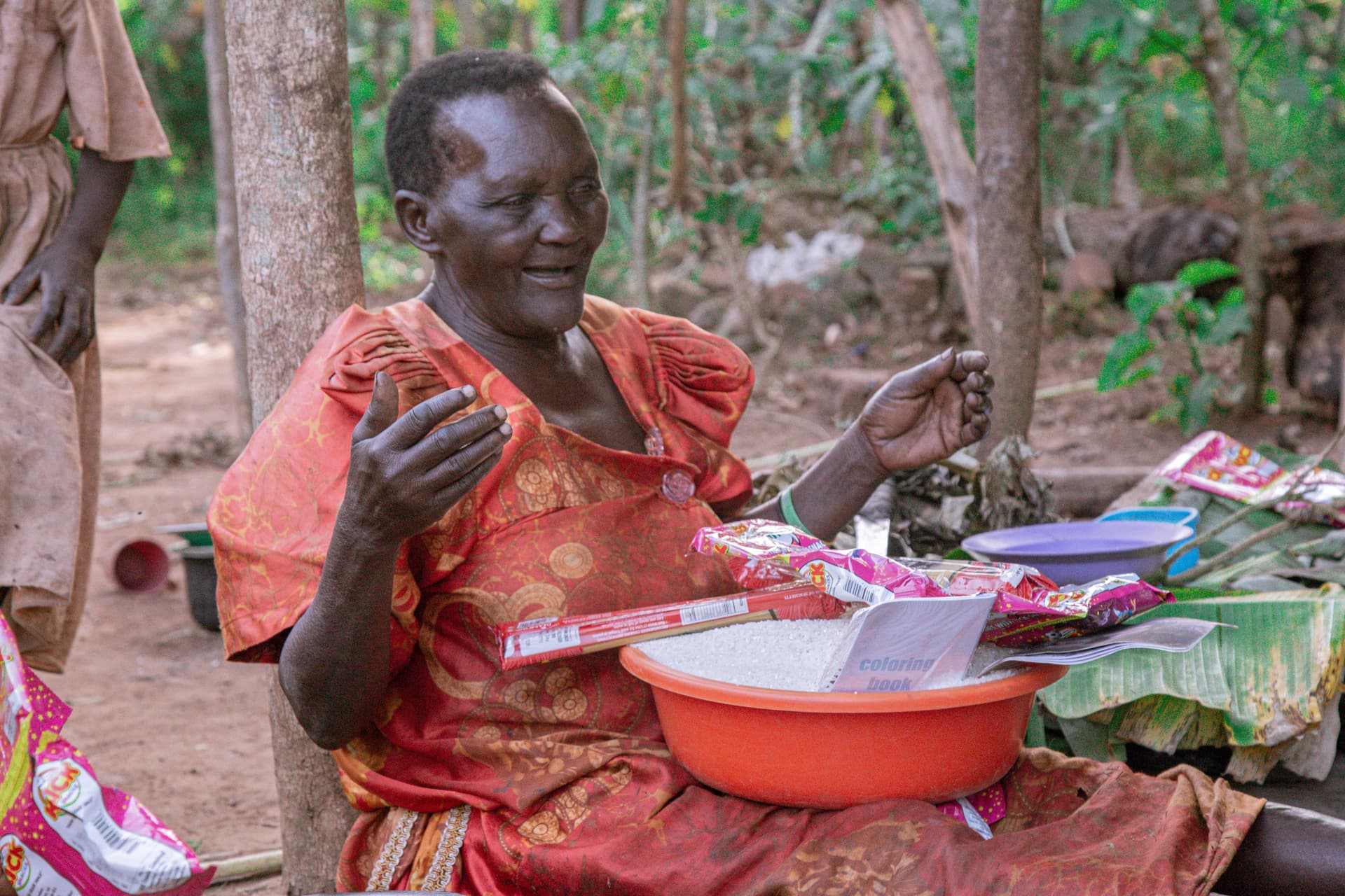 Granny Kate, an elderly woman in an orange dress, sits outdoors beside a red basin and pink food packages.