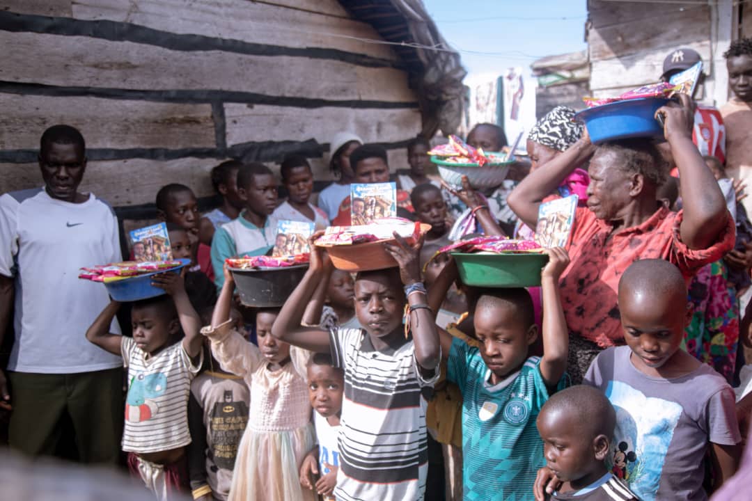 A line of women and children carrying basins of food on their heads through a village.