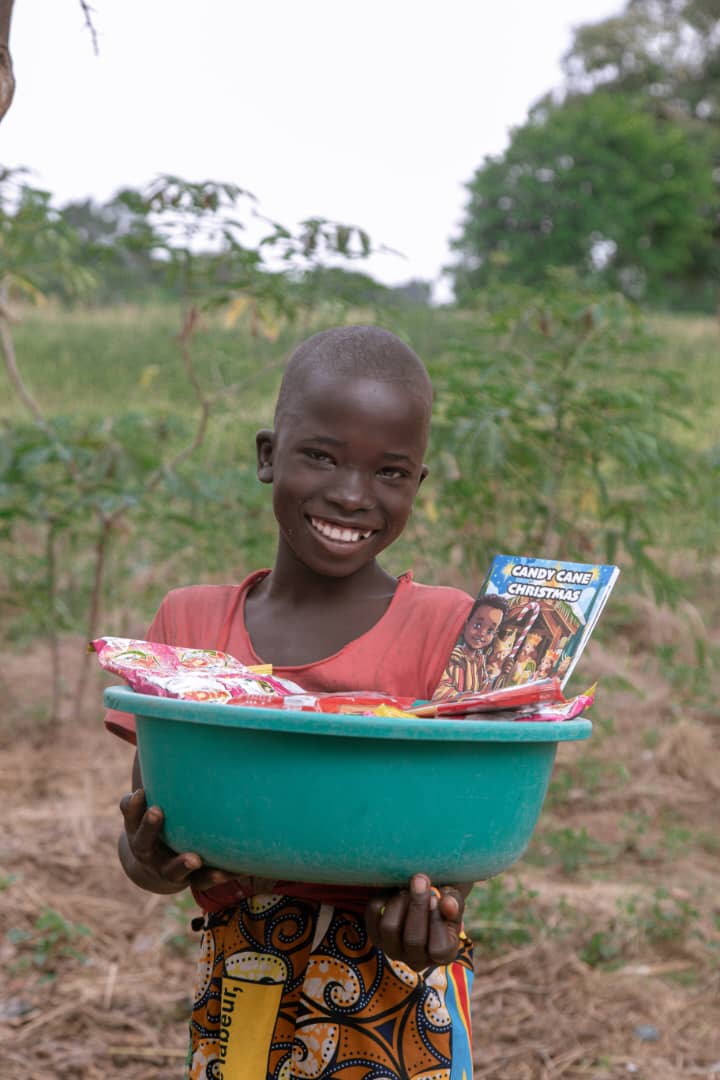 A smiling girl holds a teal basin containing food packets and a photo, standing in an open field.