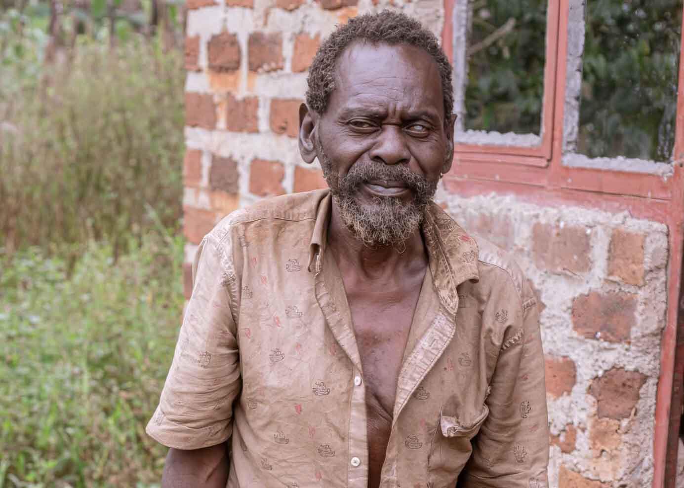 Elderly man in a tan collared shirt stands outside an unfinished brick building.